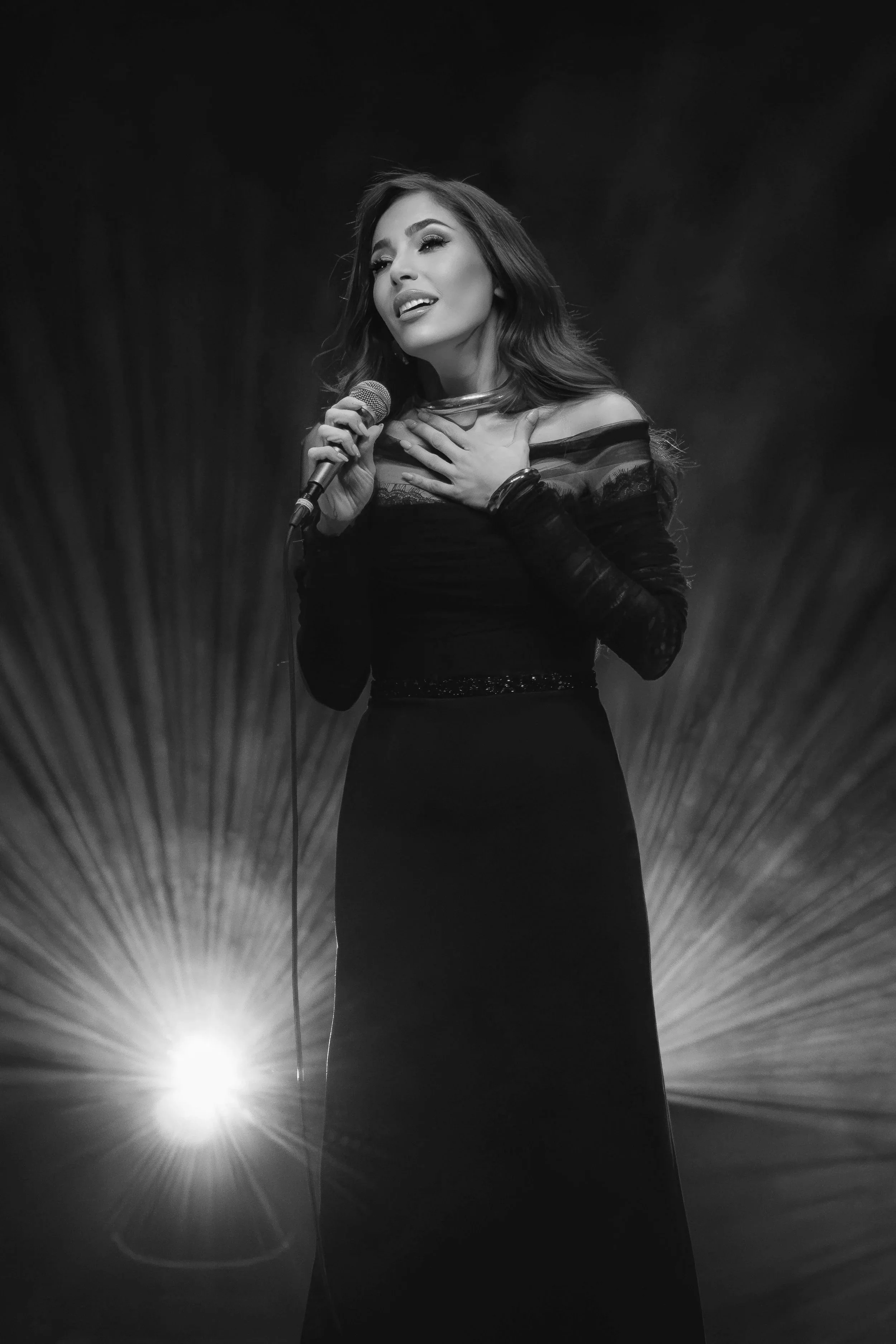 Zwart-wit foto van een vrouw die zingt met een microfoon, haar hand op haar hart en een lichte stralenbundel op de achtergrond.