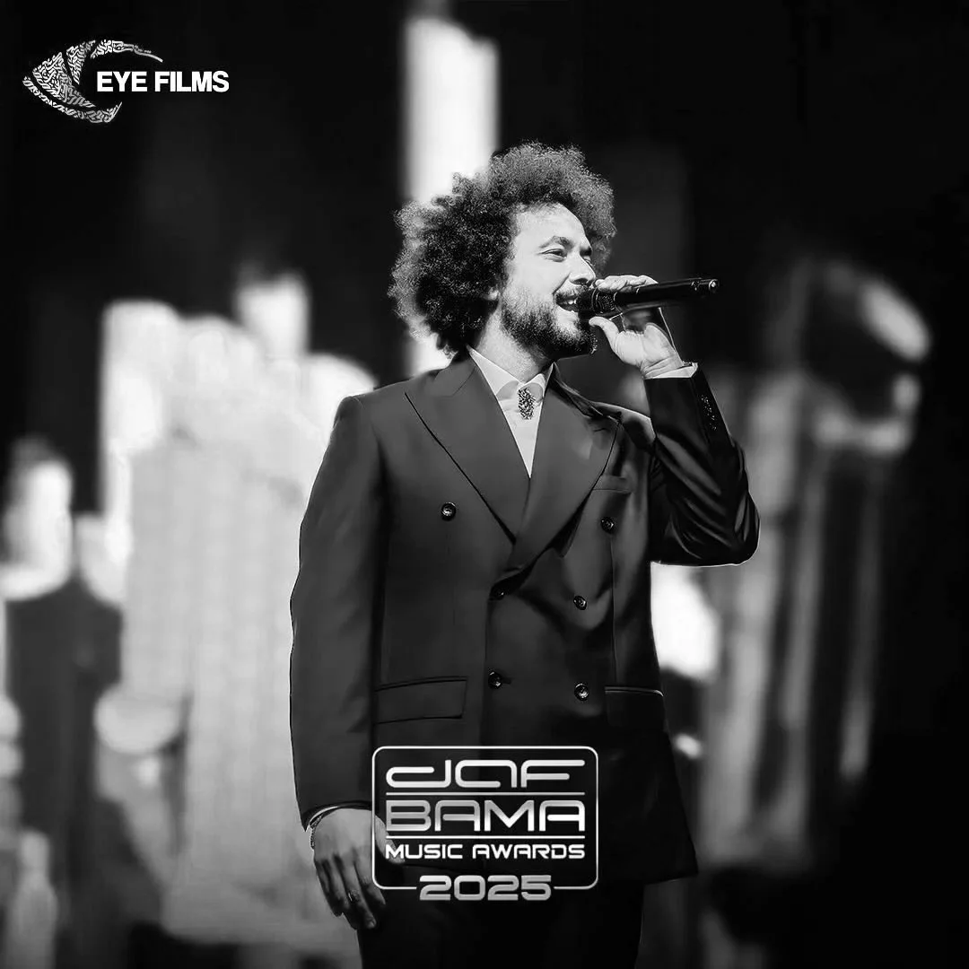 Man in zwart pak met microfoon op podium tijdens een muziekprijsuitreiking, zwart-wout foto, logo van de Grammy Awards 2025 en 'JAF BAMA MUSIC AWARDS 2025' op de afbeelding.