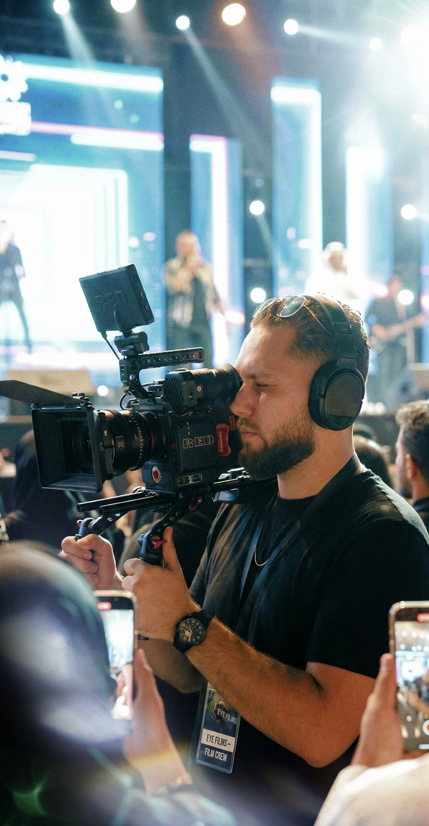Een man met koptelefoon en horloge filmt met een professionele camera in een drukke omgeving, met mensen die op de achtergrond een optreden of presentatie bijwonen.