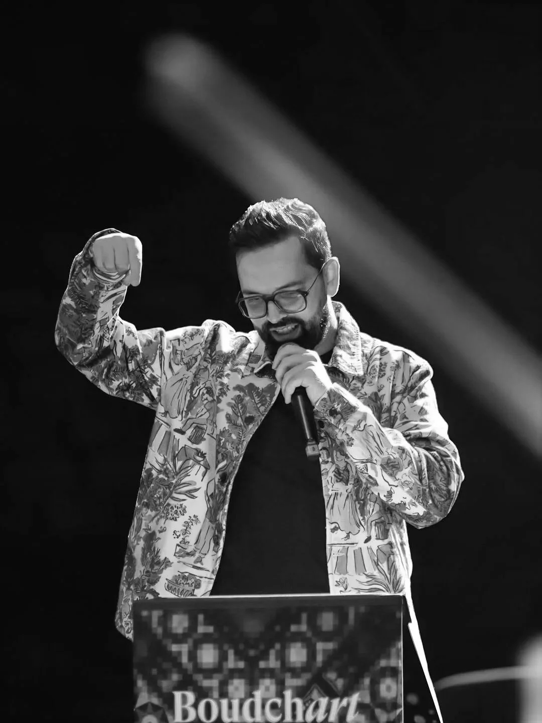 Man met bril en beard, zittend achter een DJ-achtergrond met de naam 'Boudchard', houdt een microfoon en maakt een gebaar met zijn rechterarm, in een zwart-wit foto.