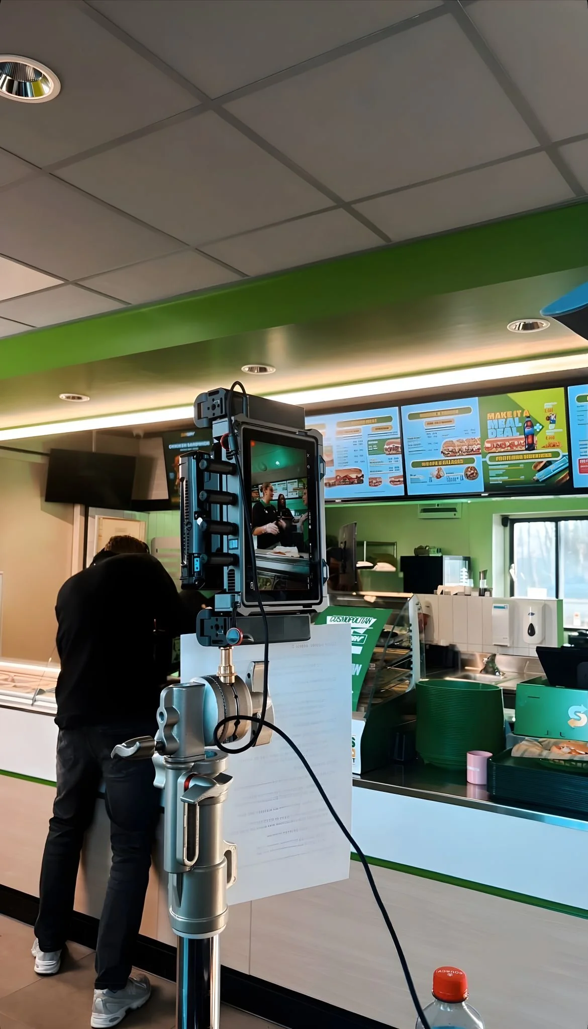 Binnen in een restaurant of fastfoodketen met een menuscherm boven de counter. Mensen wachten op hun bestelling, een robotarm is zichtbaar op de voorgrond.