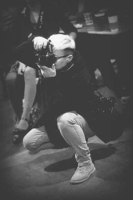 A person with light-colored hair taking a photograph while crouching.