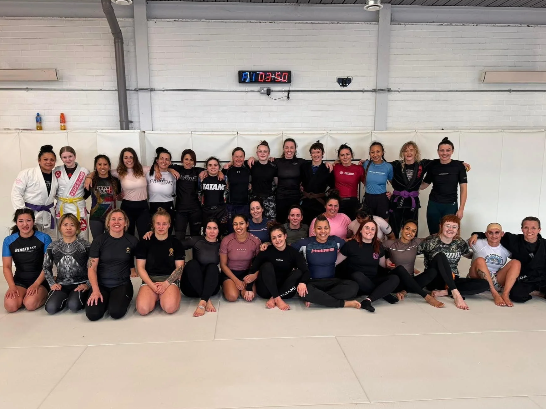 Group of women posing together in martial arts uniforms and athletic wear inside a training facility with a white brick wall and digital clock on the wall.