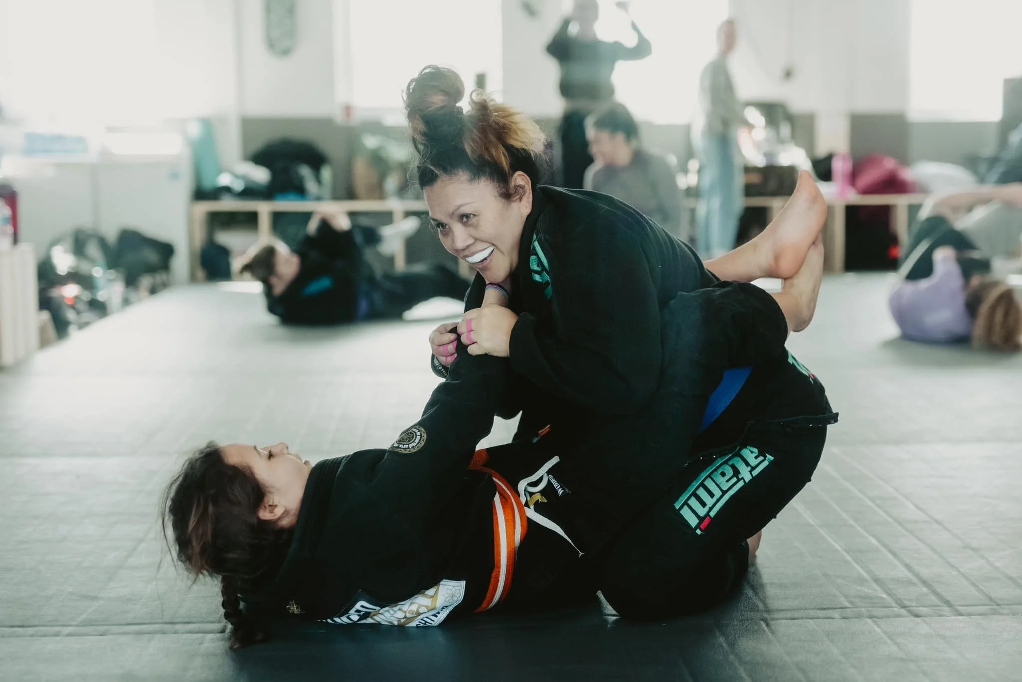 Two women practicing Brazilian Jiu-Jitsu on a mat in a gym, with one woman on top applying a submission hold on the other woman.