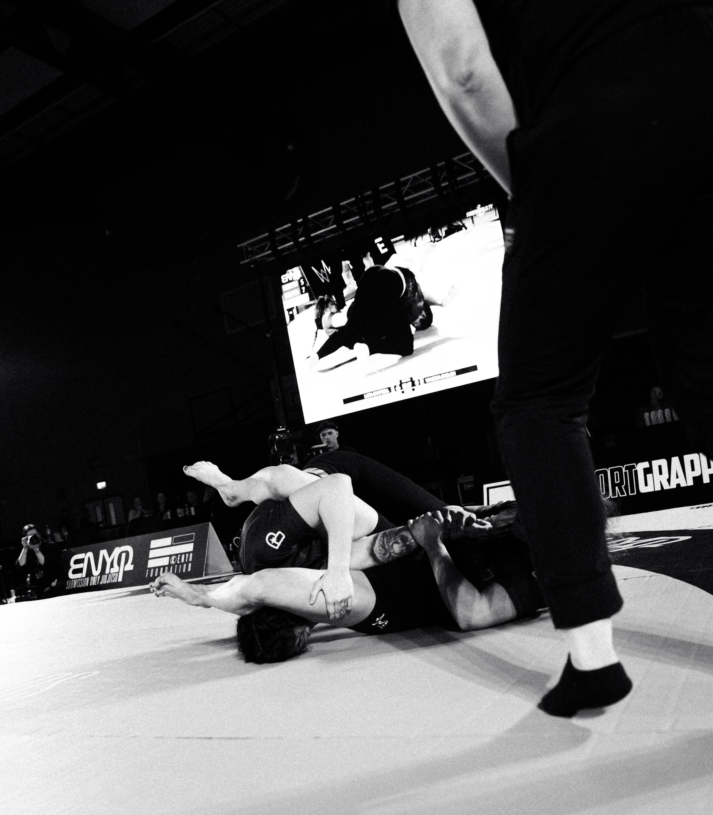 Two martial artists in a competitive Brazilian Jiu-Jitsu match, with one on top and the other on the ground, both wearing rash guards and shorts, in an indoor gym with a large screen displaying advertisements in the background.