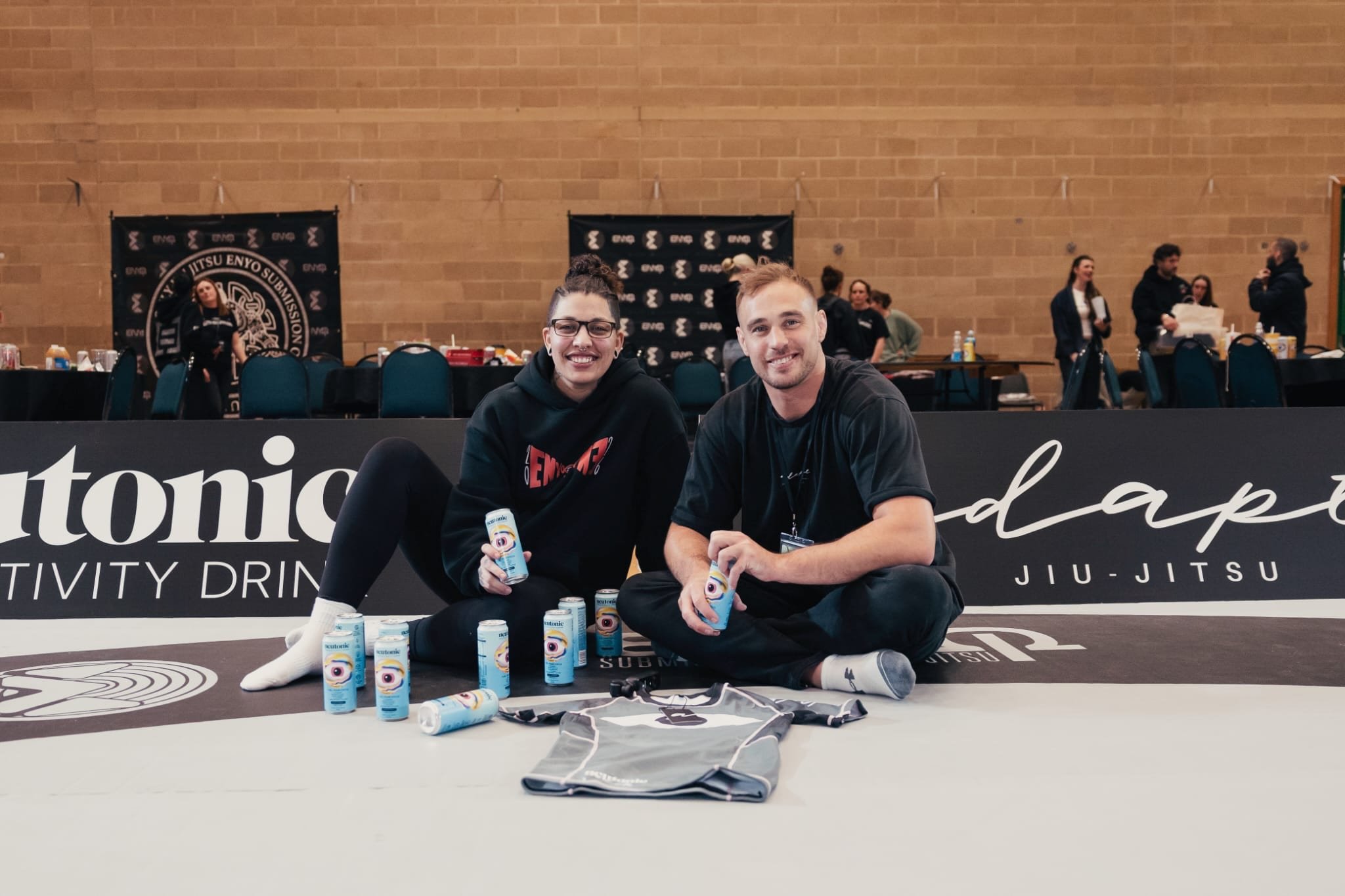 Two smiling young adults sitting on the floor in front of a black banner, holding cans of Kudutonic beverage, surrounded by additional cans, with a gymnasium background and people in the background.