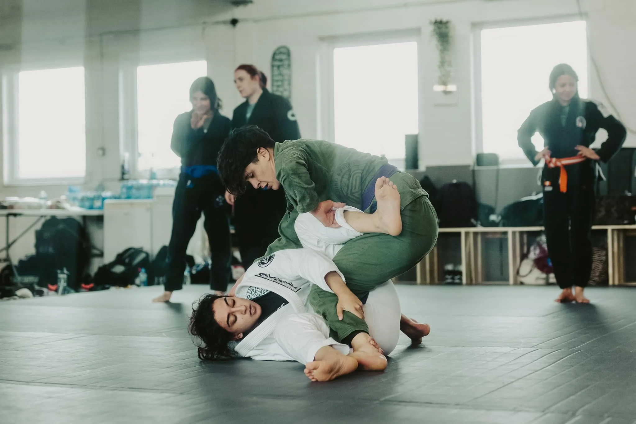 Two women practicing Brazilian Jiu-Jitsu on the mat with one woman in a white gi on the ground and the other woman in a green gi on top, while others watch in the background.