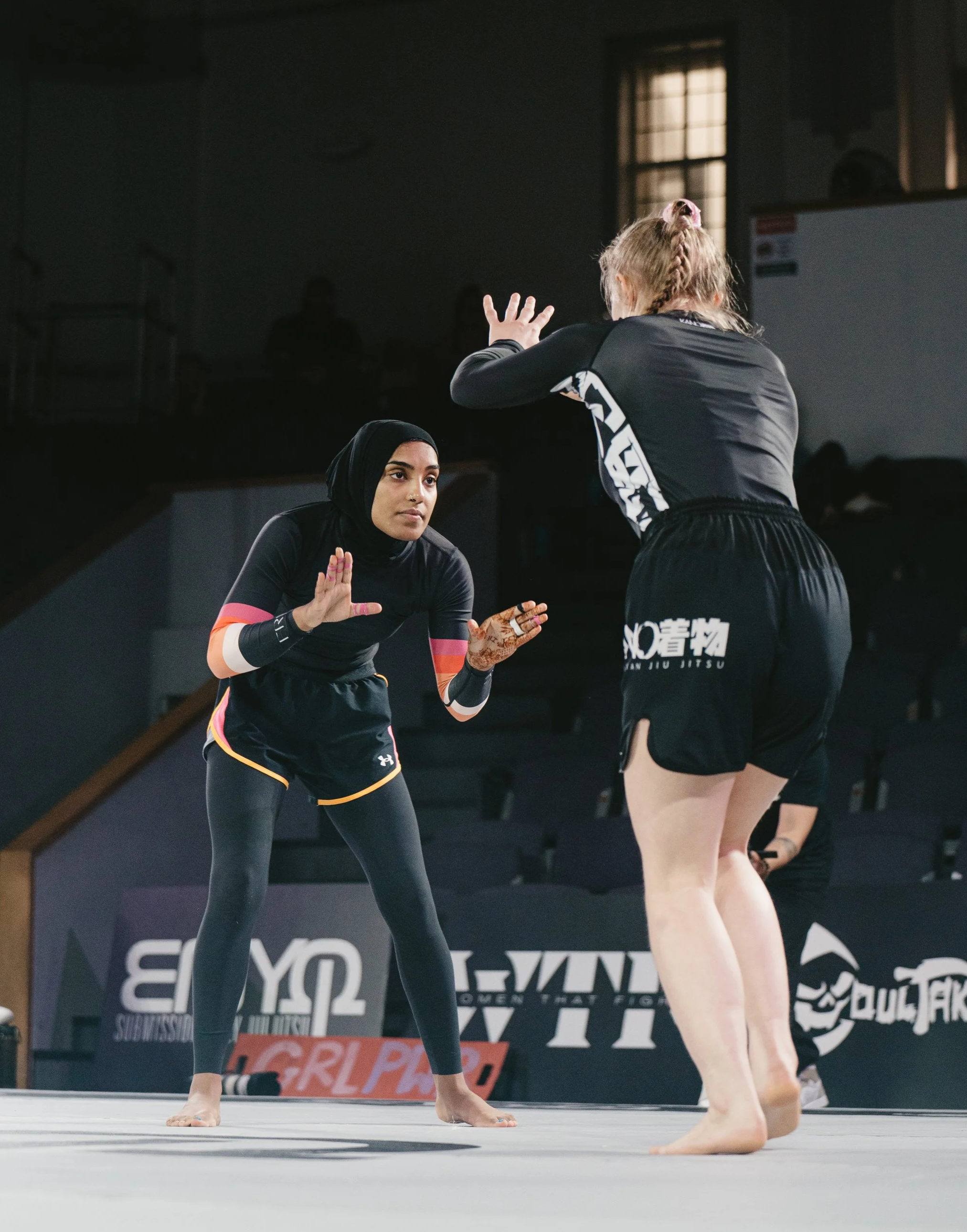 Two female athletes practicing Brazilian Jiu Jitsu during a match on a mat indoors, with dark background, banners, and spectators in the background.