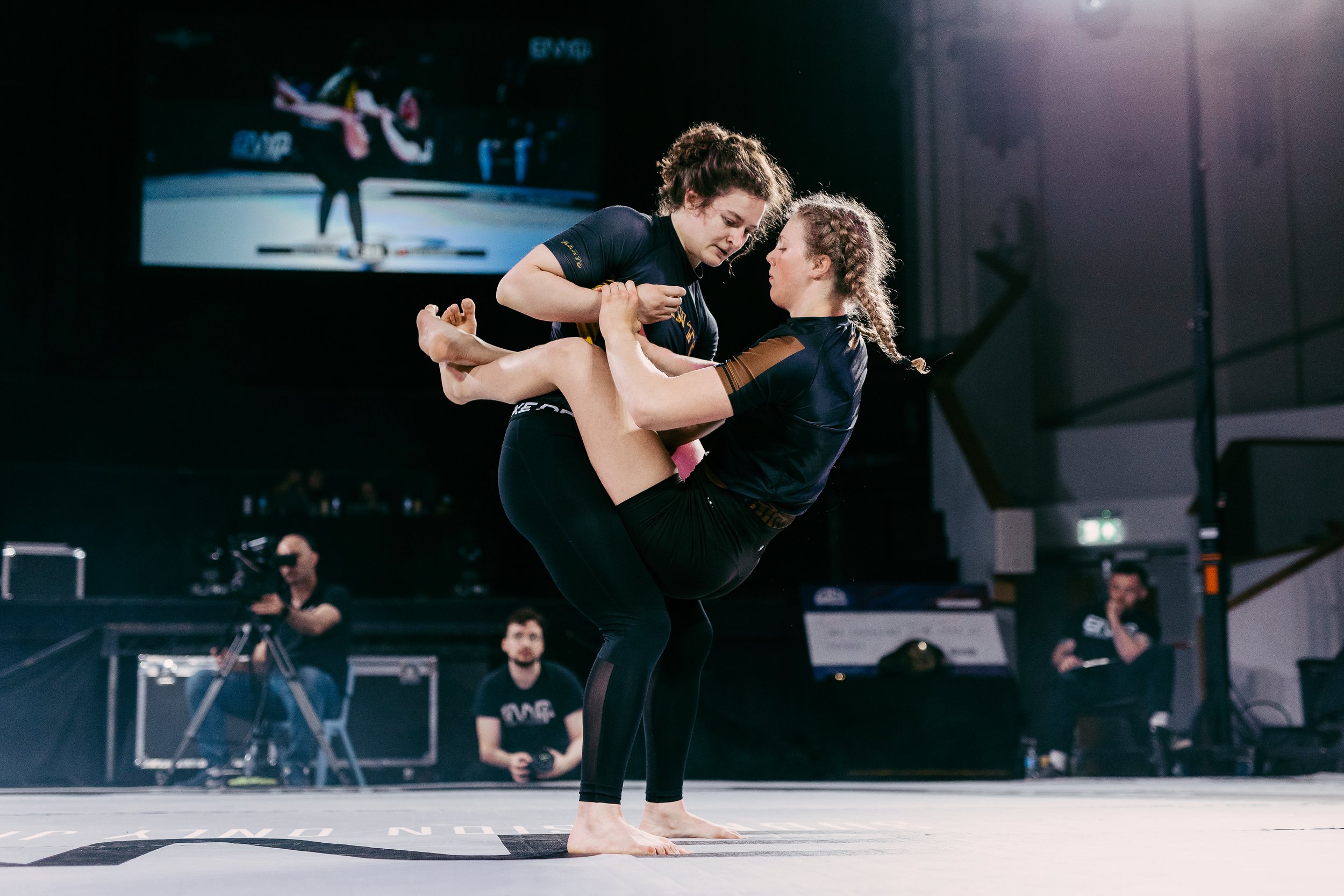 Two women practicing yoga or martial arts on a mat in a gym, with one woman supporting the other who is holding a challenging pose. Several people are watching in the background, some with cameras, and a large screen shows a similar scene.