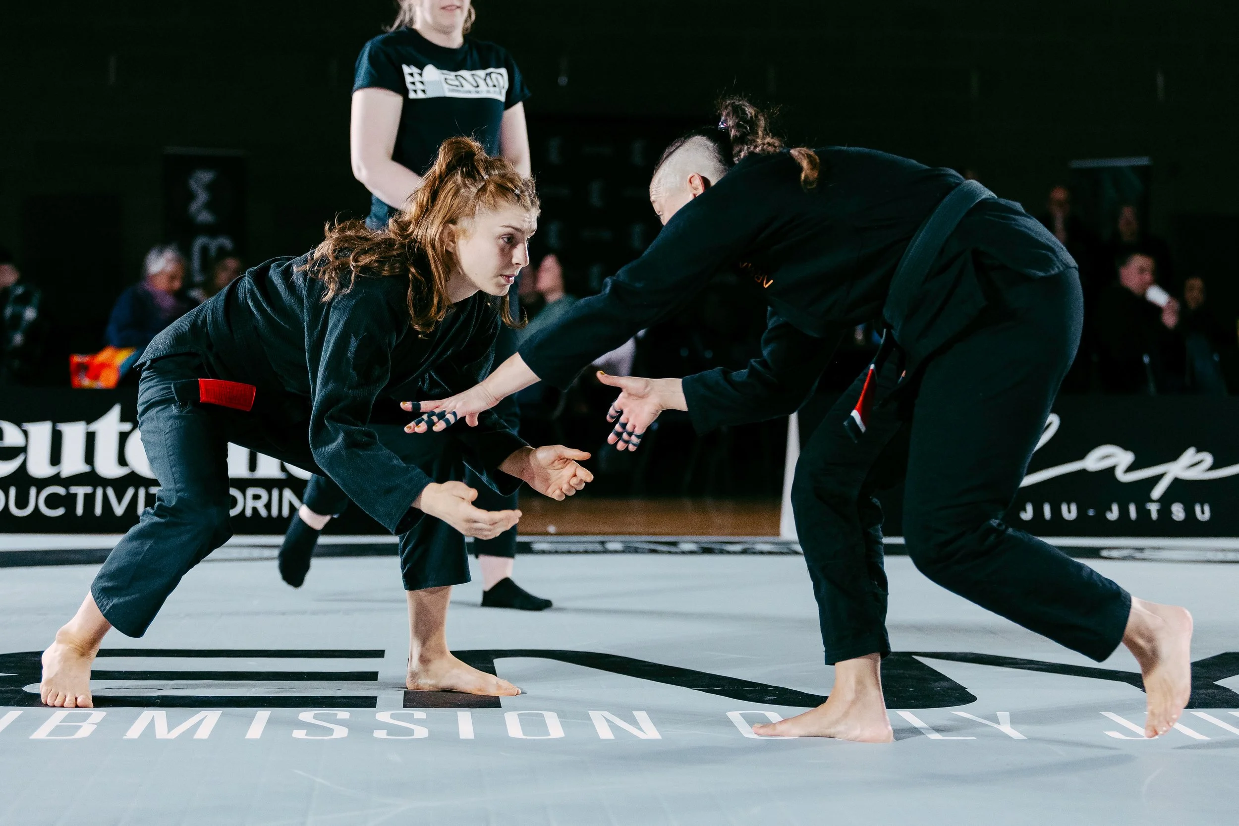 Two women practicing Brazilian Jiu-Jitsu on a mat, facing each other in a grappling stance, with a referee or coach observing nearby.