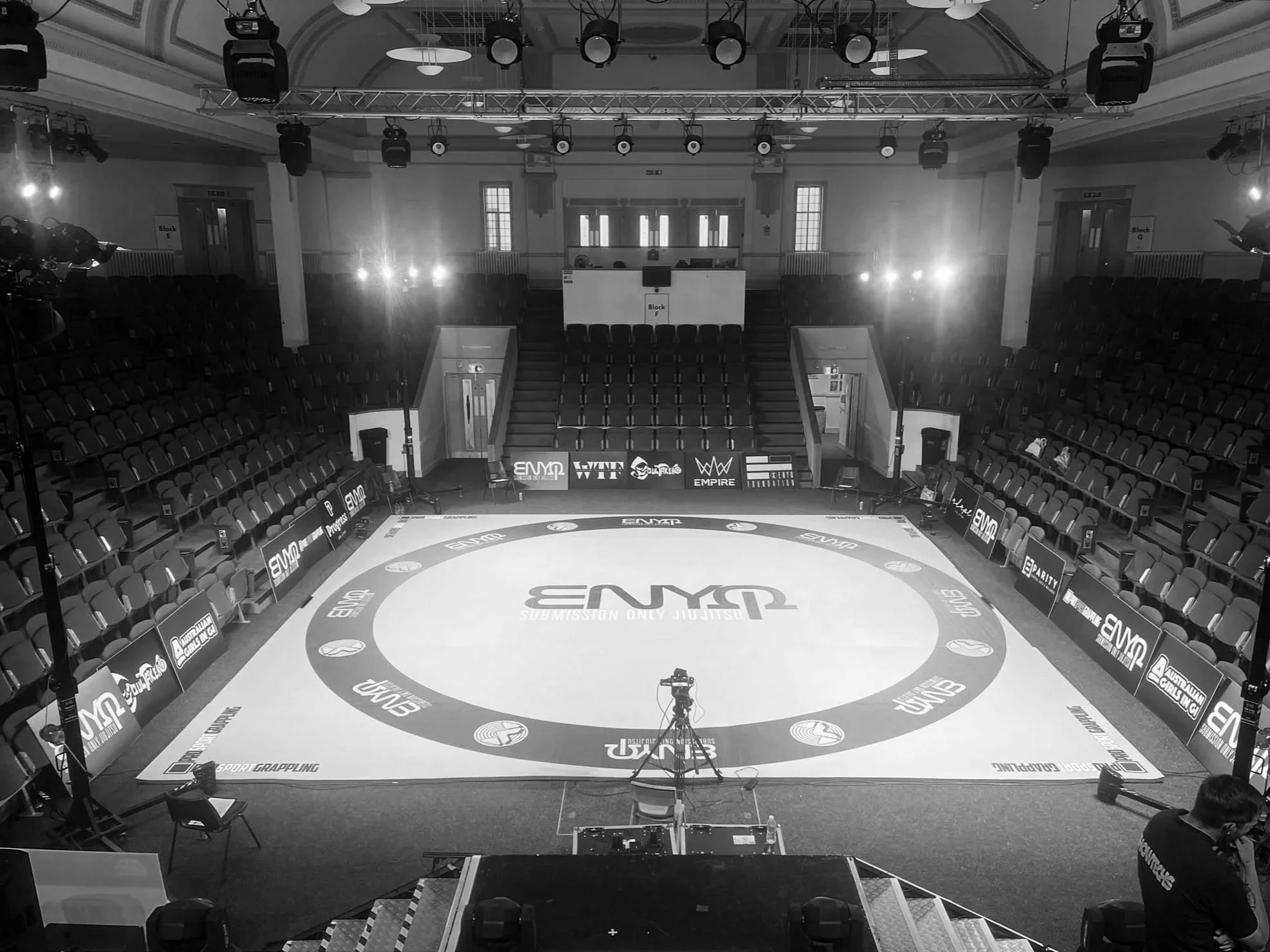 An empty indoor sports arena set up for a grappling or martial arts event. The circular mat has the logo 'ENYO' and is surrounded by chairs and banners. There is a camera on a tripod in front of the mat, and lighting equipment is suspended from the ceiling.