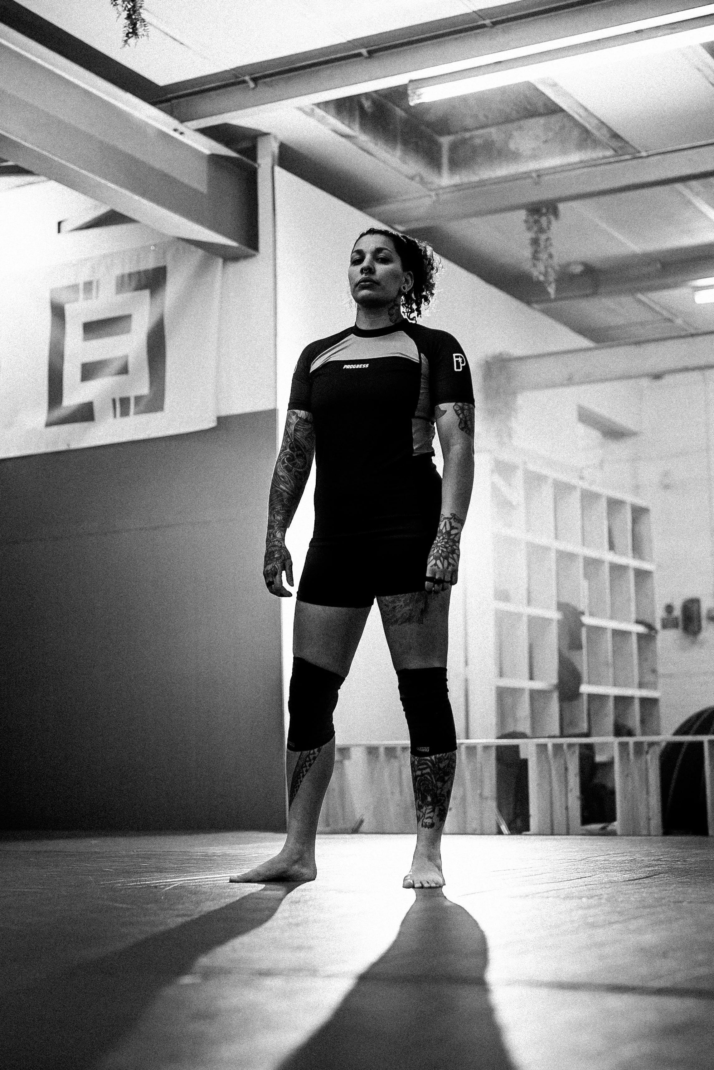A woman standing on a martial arts training mat in a gym, with tattoos on her arms and legs, wearing a black athletic outfit, looking confident, with gym equipment in the background.