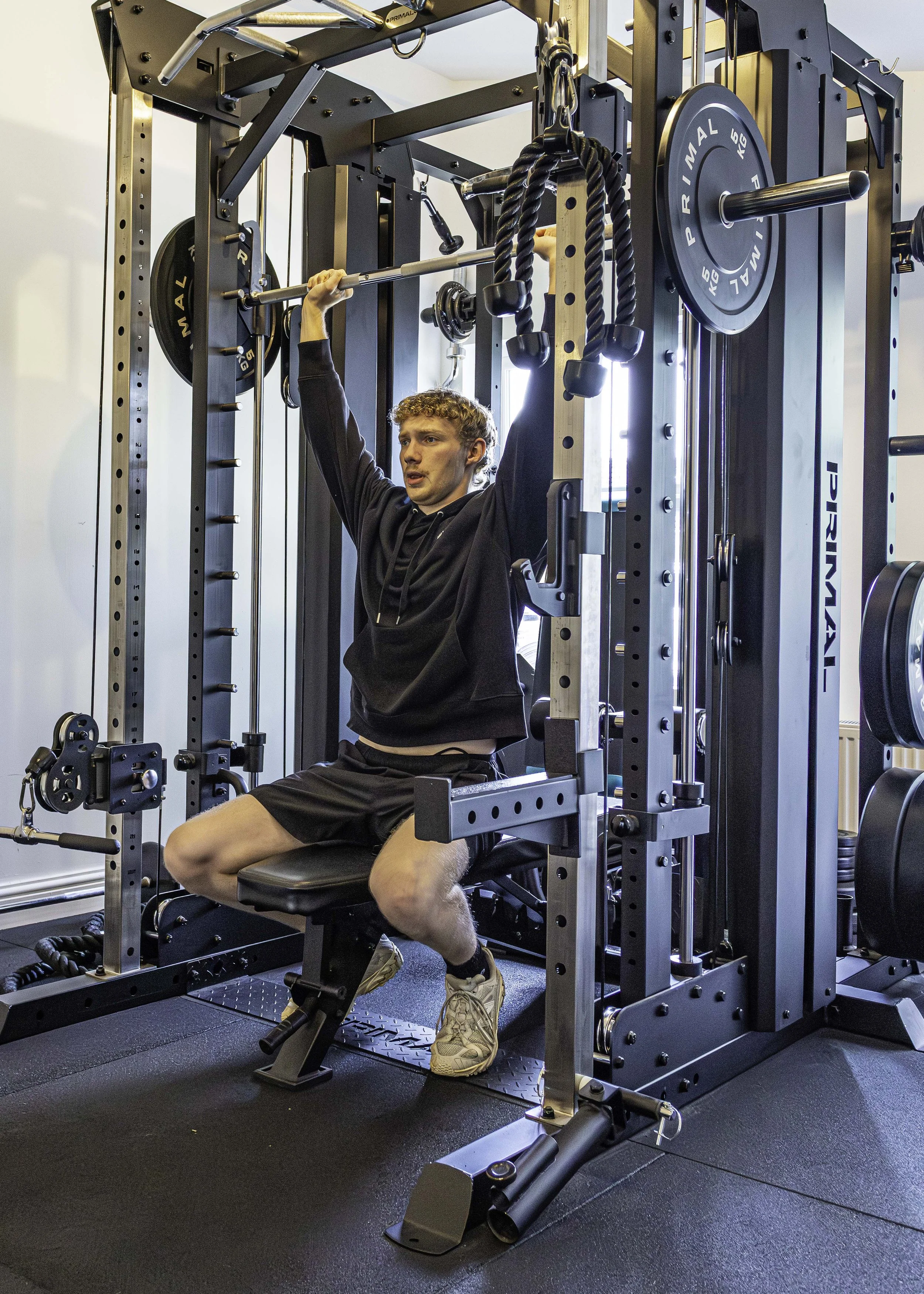 A young man with curly hair in a black hoodie and shorts is exercising on a seated smith machine in a gym. He is lifting a barbell with weights. The gym has black flooring and workout equipment.