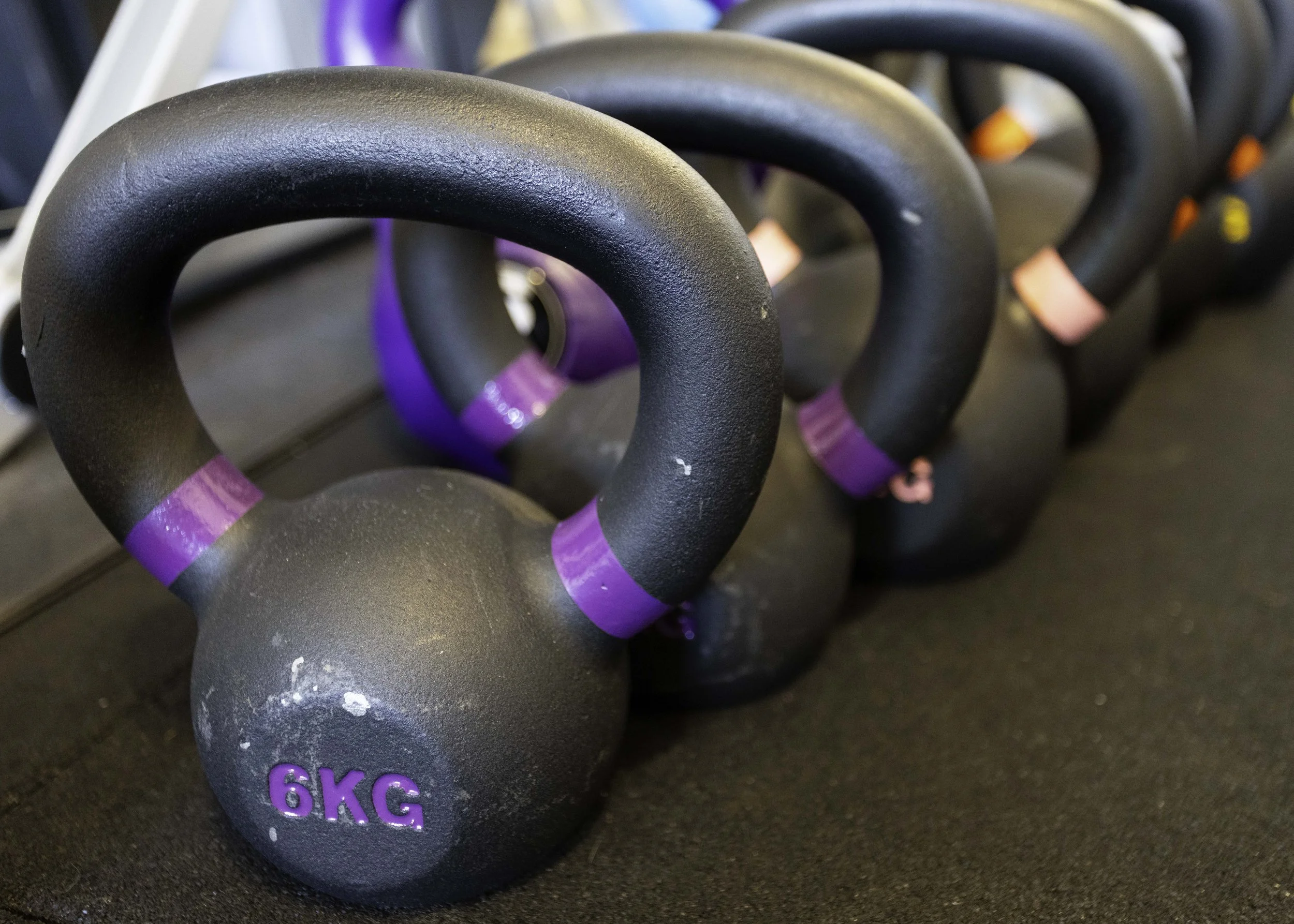 Close-up of black kettlebells arranged in a line on a dark exercise mat, with purple and pink markings indicating weight, including one labeled "6Kg."