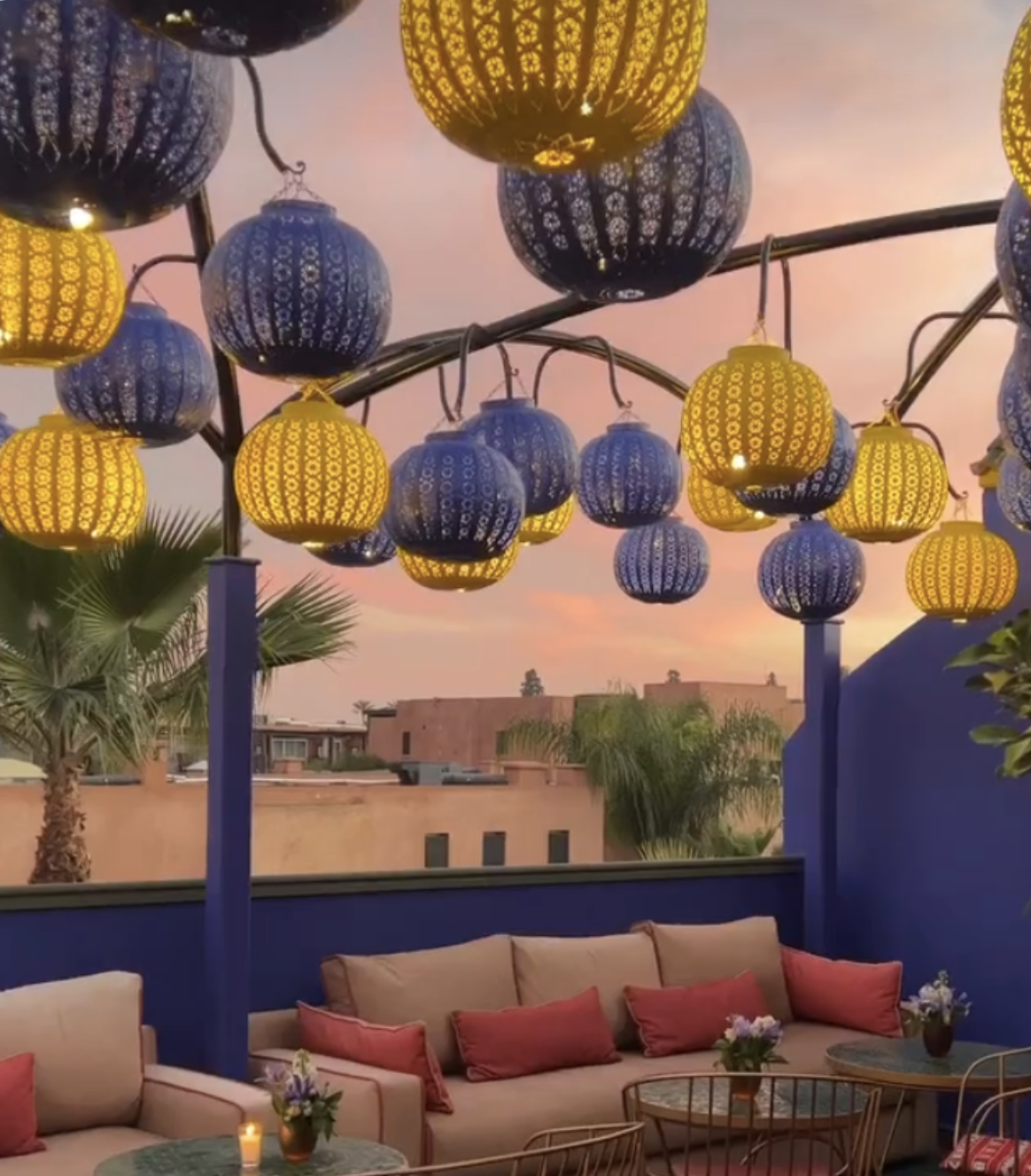 An outdoor terrace event design inspired by Le Jardin Majorelle, showcasing an overhead canopy of hanging blue and yellow fretwork lanterns above modern patio sofas with red accent cushions at sunset.
