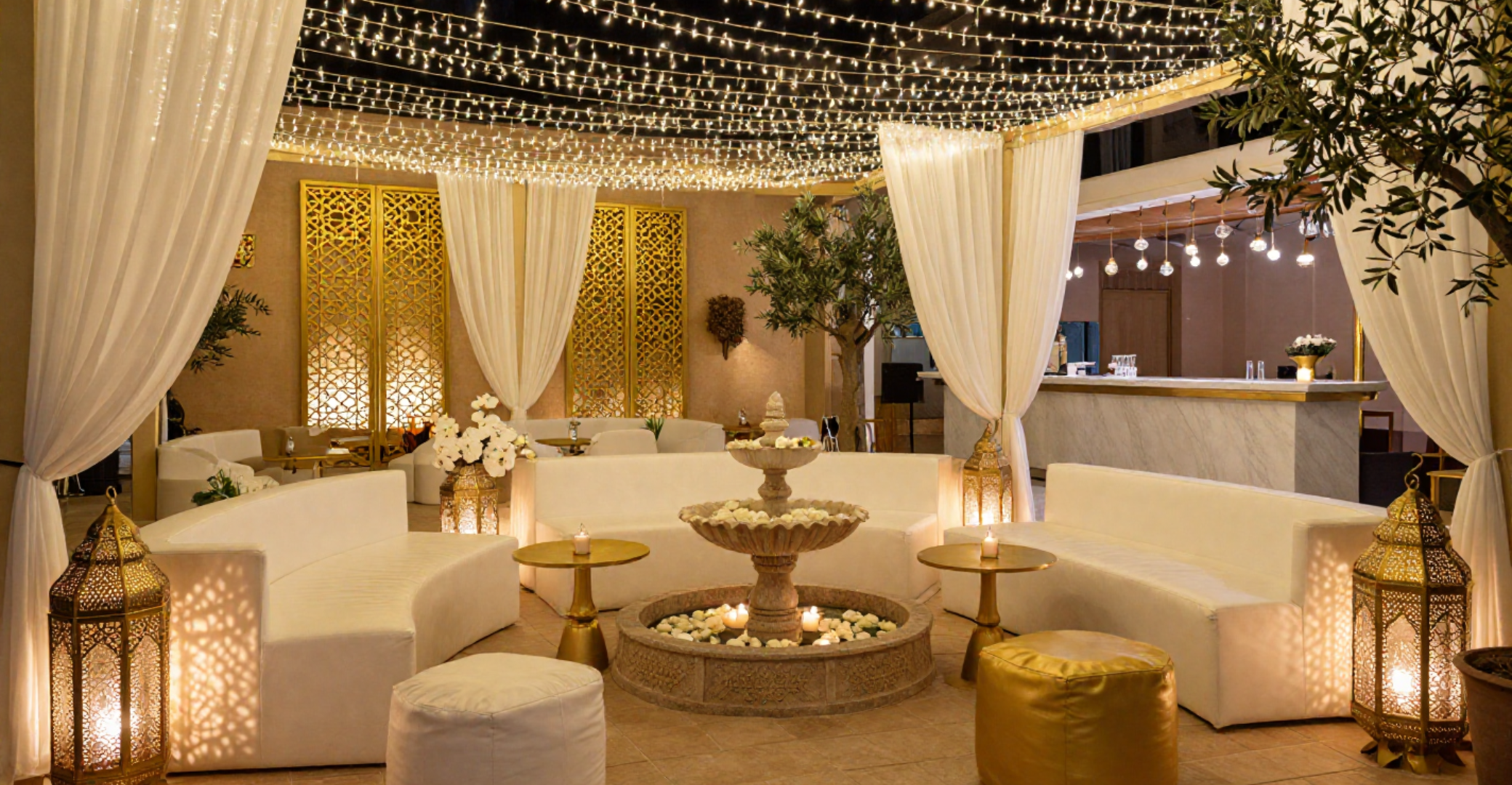 An elegantly decorated indoor lounge area with white curved banquette seating, small tables, and lanterns. The ceiling is adorned with string lights, and there are gold decorative screens and planters with trees and flowers. The ambiance is warm and inviting, suggesting a setting for a celebration or special event.