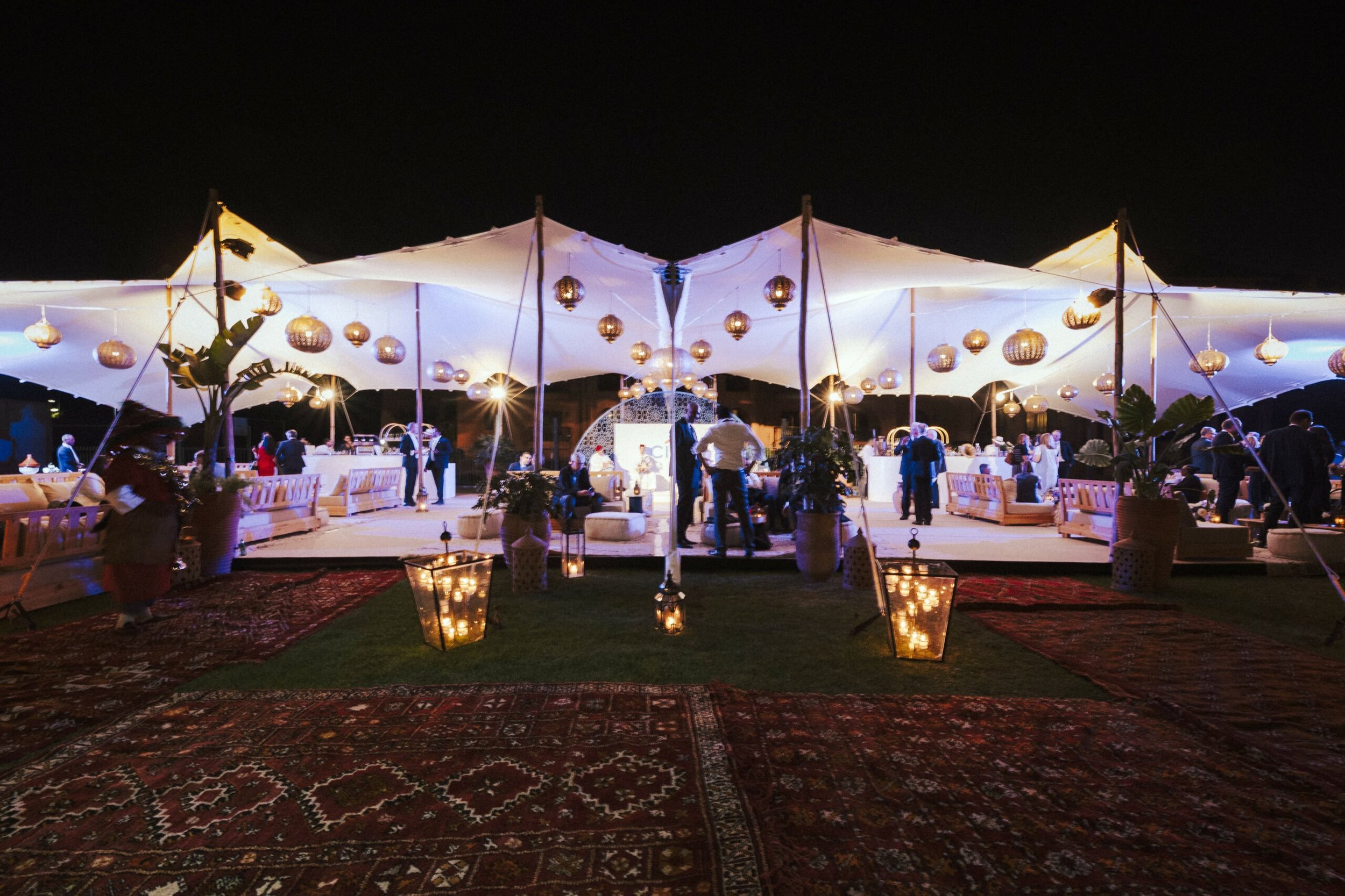 Night scene of an outdoor event under a large white canopy decorated with hanging lanterns, with people standing and sitting, and a patterned red carpet on the ground.