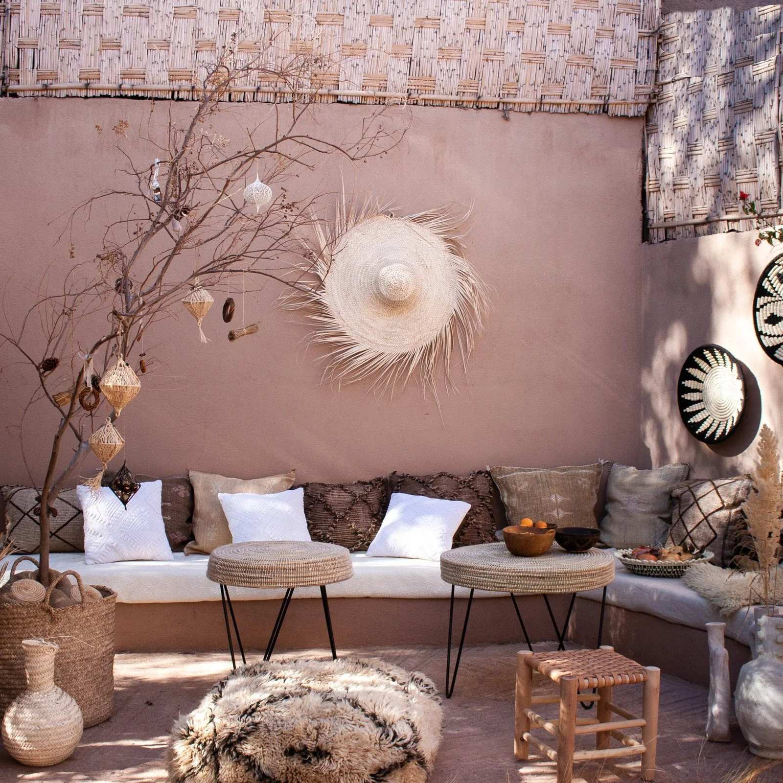 Modern boho chic Moroccan tea lounge setting against a terracotta wall featuring a low plaster built-in sofa, woven straw tables, neutral patterned throw pillows, and dried botanical branch accents.
