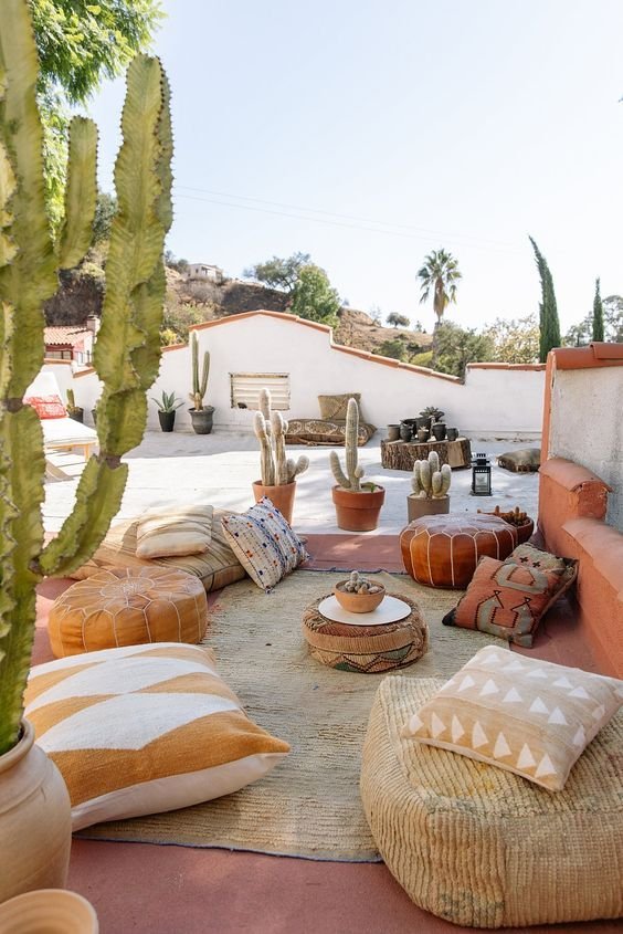 Outdoor terrace with desert-themed decor, including large cacti potted in terracotta, cushions and poufs on a rug, and a rustic white wall with orange trim in the background. Hills and palm trees are visible in the distance.