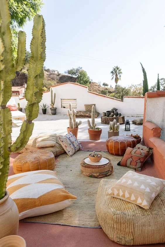 A cozy bohemian-style outdoor balcony floor seating arrangement with light-textured jute rugs, oversized patterned geometric pillows, and a tan leather pouf.