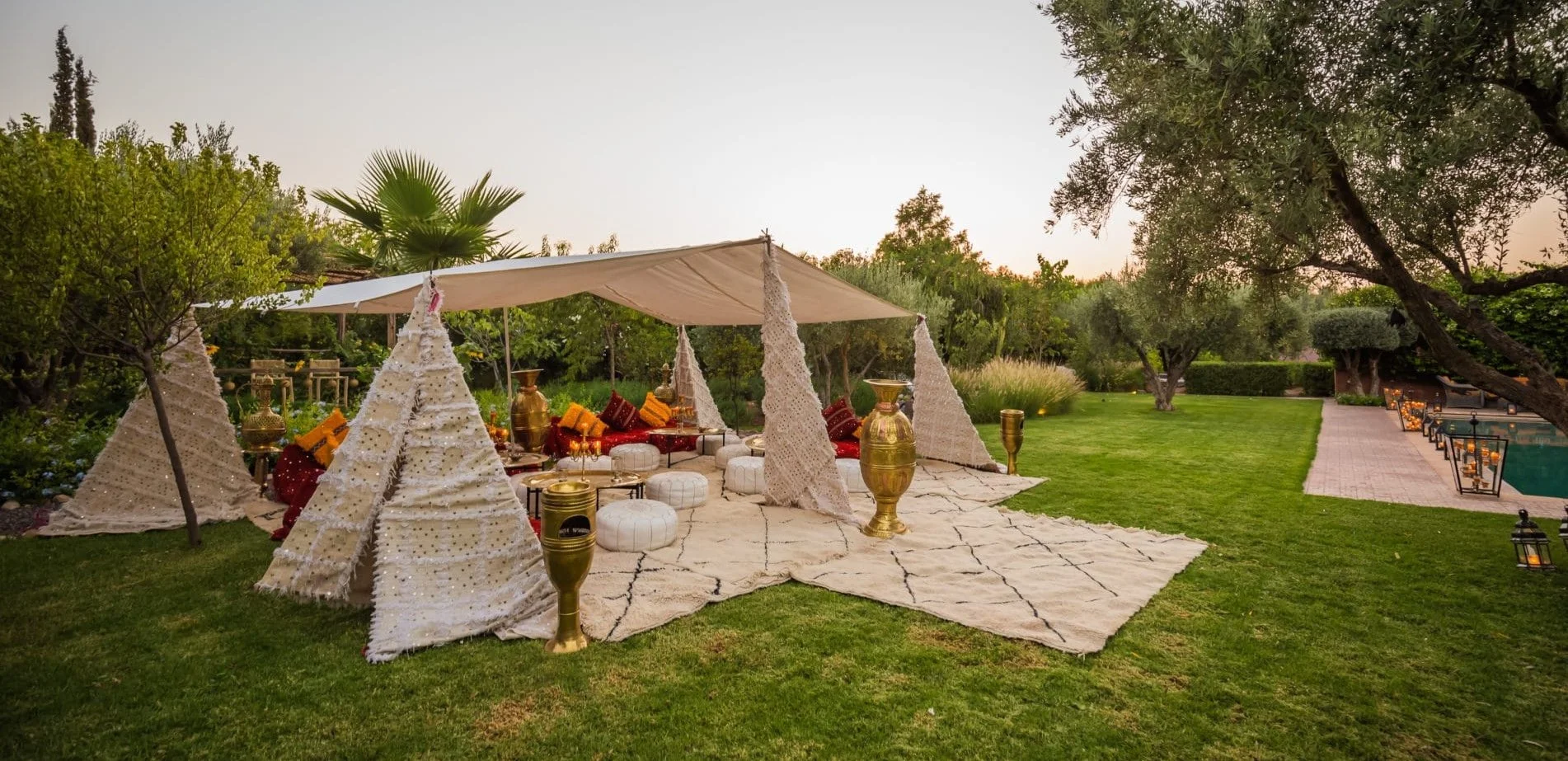An elegant outdoor backyard event lounge styled with white woven sequined Moroccan wedding blankets draped over a canopy tent structure, premium textured white floor cushions, large polished gold statement urns, and layered cream area carpets.
