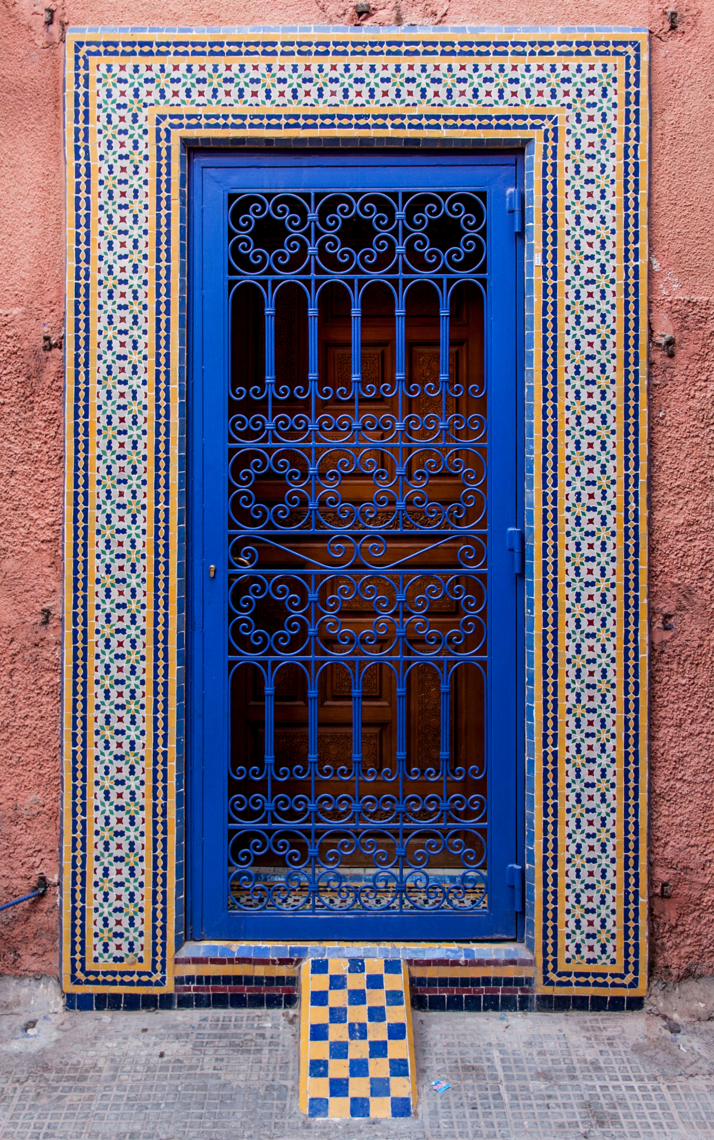 A striking authentic Moroccan door backdrop painted in rich Majorelle blue with intricate black wrought iron scrollwork, framed by traditional hand-cut mosaic tiles in geometric blue, white, and yellow patterns.