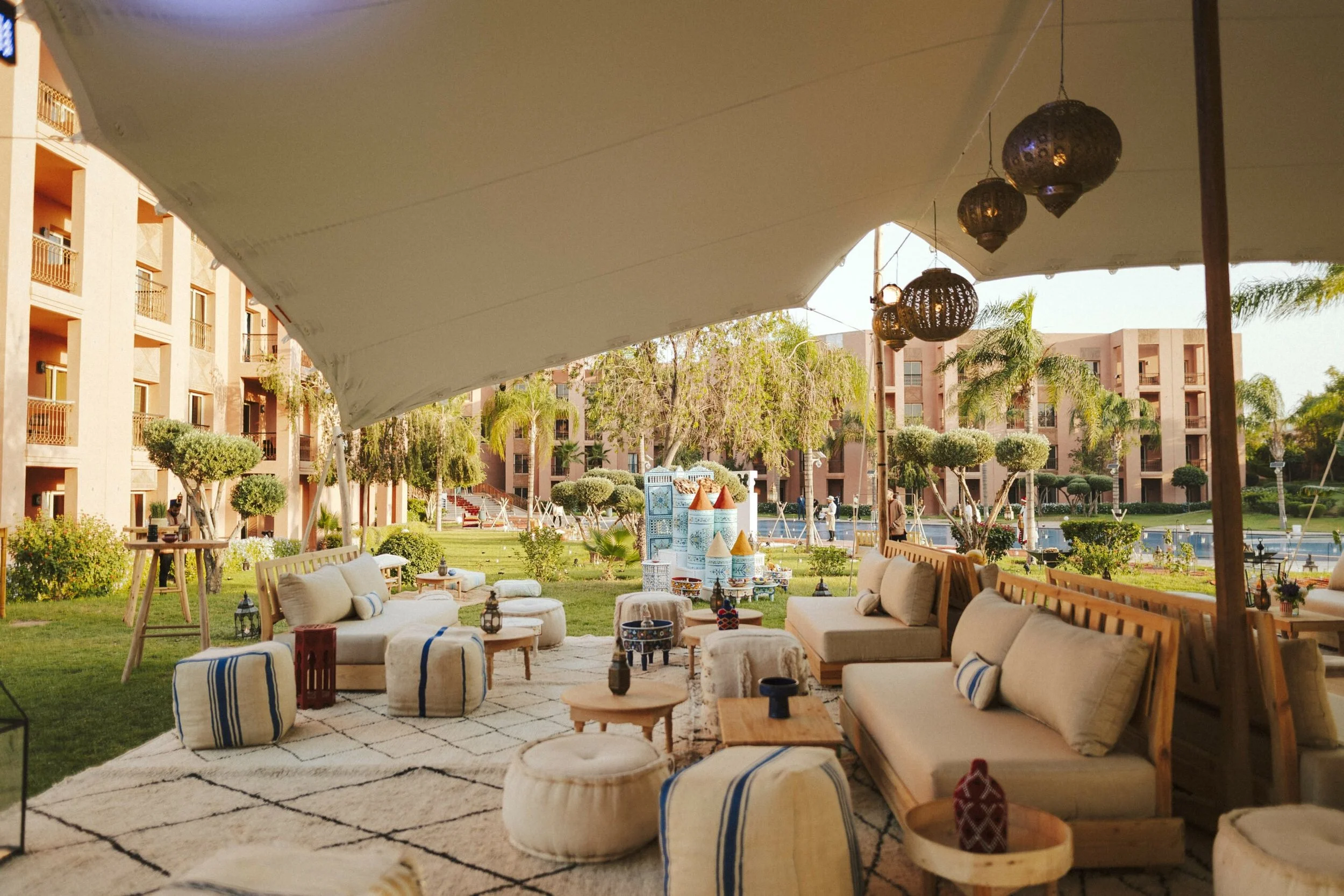 Upscale outdoor event lounge under a white shade canopy featuring modern neutral sofas, woven cream floor poufs, low wooden tables, and hanging openwork brass lanterns.