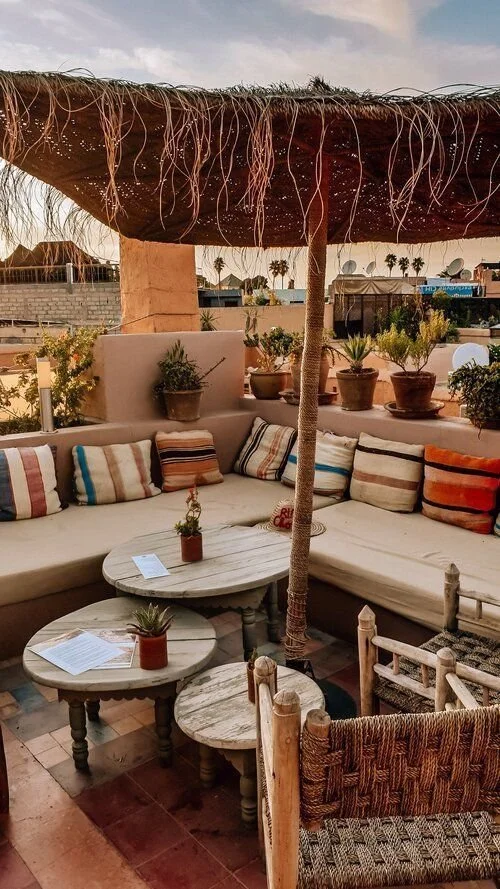 Outdoor patio with cushioned seating, striped pillows, potted plants, and a thatched umbrella in a warm, sunny setting.