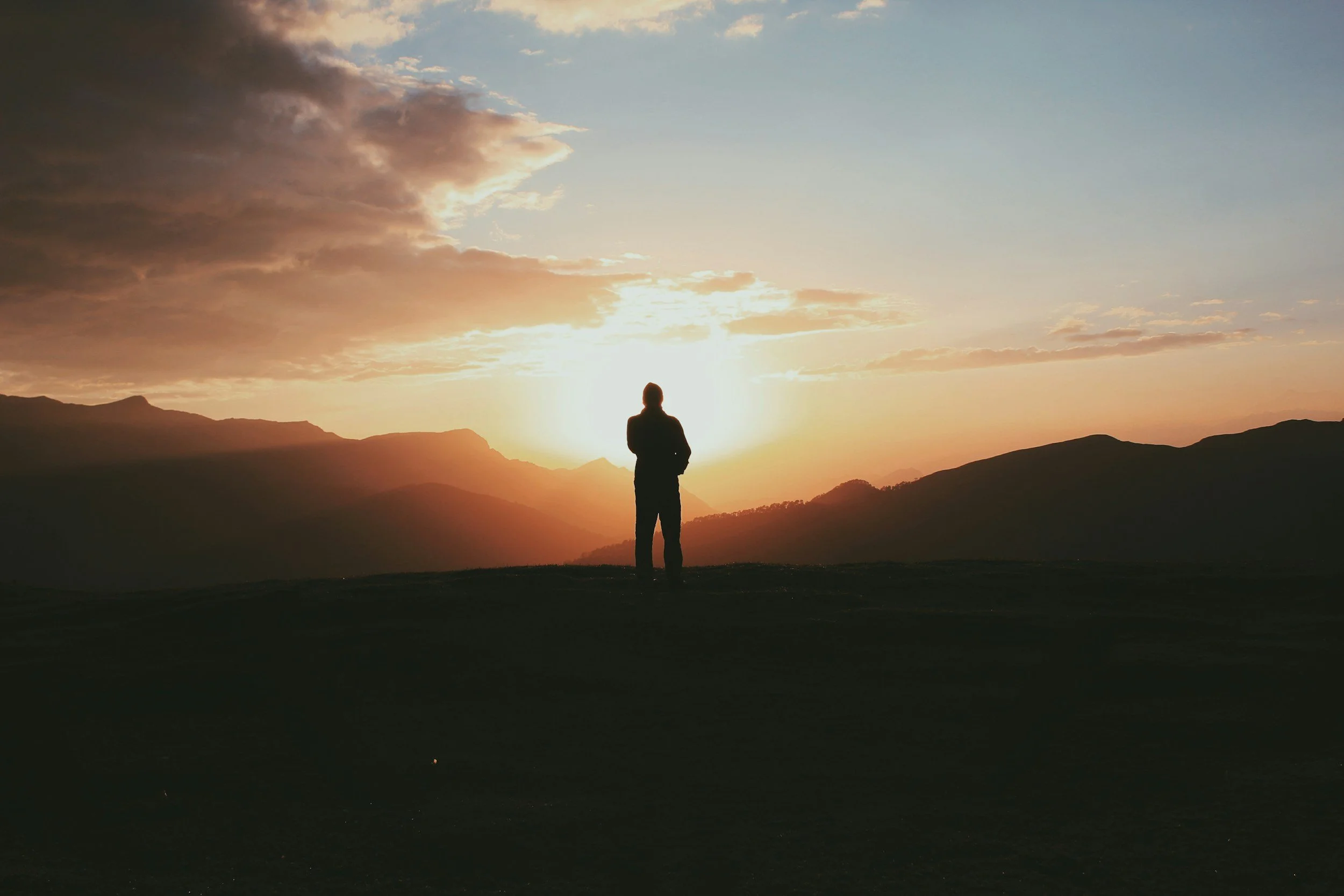 Person standing on a hill at sunset, overlooking mountains with a partly cloudy sky.