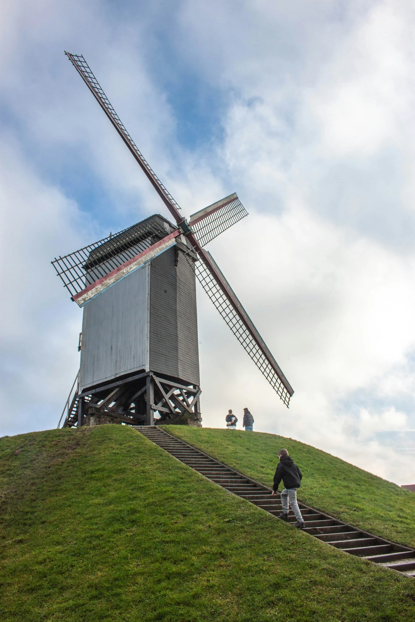 A historic wooden windmill stands on a grassy hill with a staircase leading up to it. Two people are seen near the windmill and a person is walking up the stairs.
