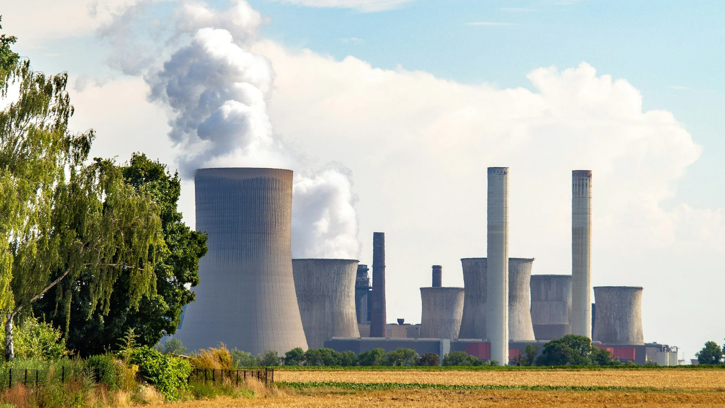 Power plant with multiple cooling towers emitting smoke or steam, surrounded by trees and fields.