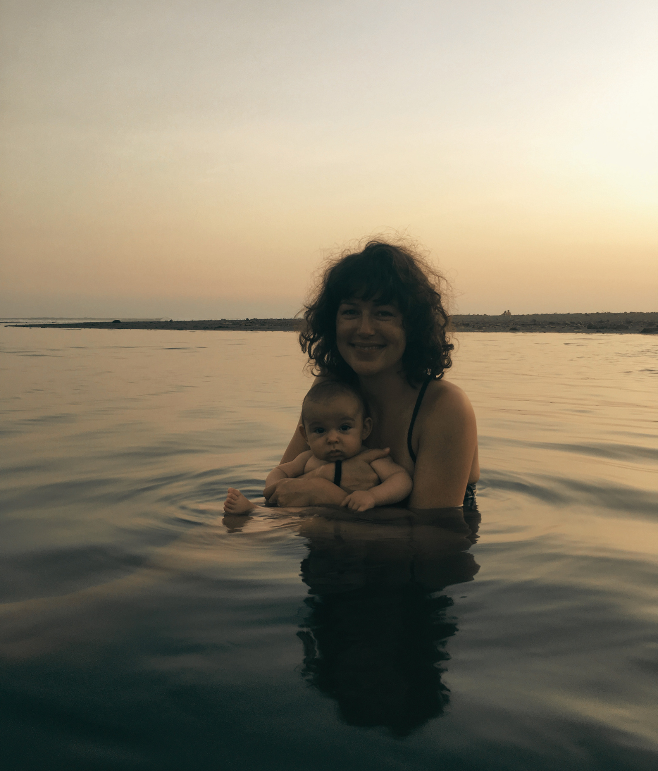 A woman with curly hair smiling while holding a baby in a lake at sunset.