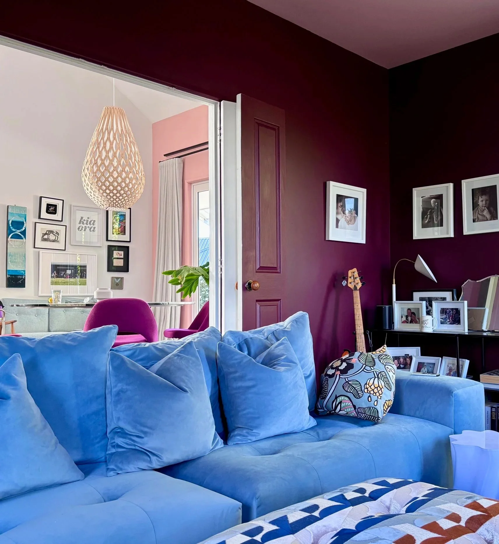 Living room with a blue couch and an upholstered armchair with a floral pattern. Behind the blue couch, there are pillows. A side table with framed photos, a mug, and other items. On the wall, there are framed photos and artwork. A guitar is leaning against the wall near a window with curtains. A white floor lamp is also visible.
