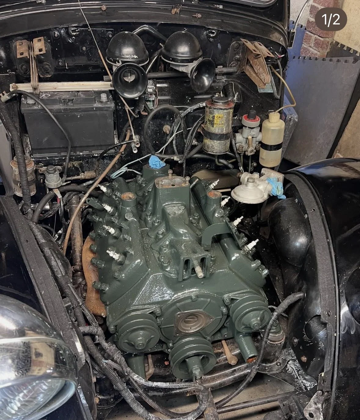 An engine bay of a vintage car, with the engine removed and replaced with a large, green-painted engine block, surrounded by various car parts, wires, and tools.