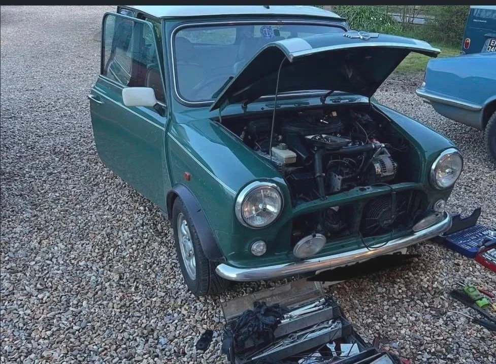 A green vintage Mini Cooper with its hood open, parked on a gravel surface, with tools and parts laying nearby, indicating repair or maintenance work.