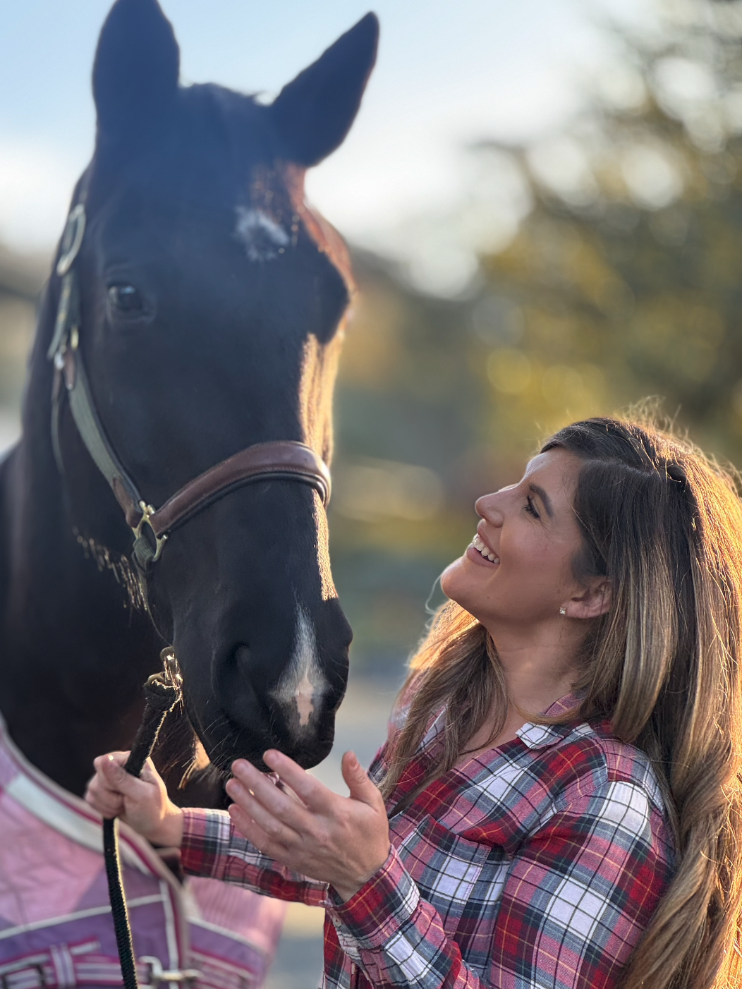 A woman smiling and looking at a black horse, holding its bridle, outdoors during sunset.