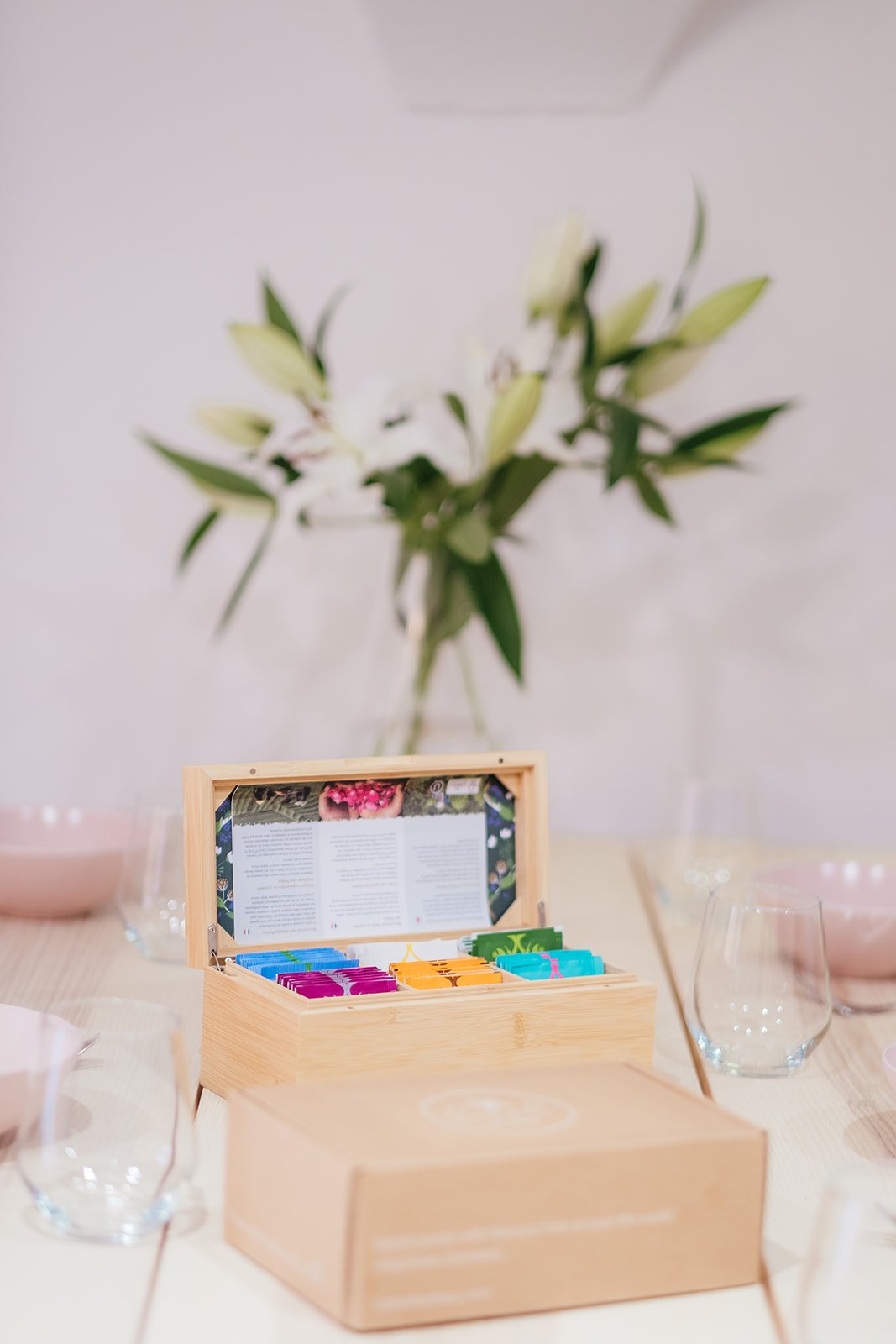 A wooden box containing tea or other small packets sits on a table with glasses around it, with a vase of white flowers in the background.