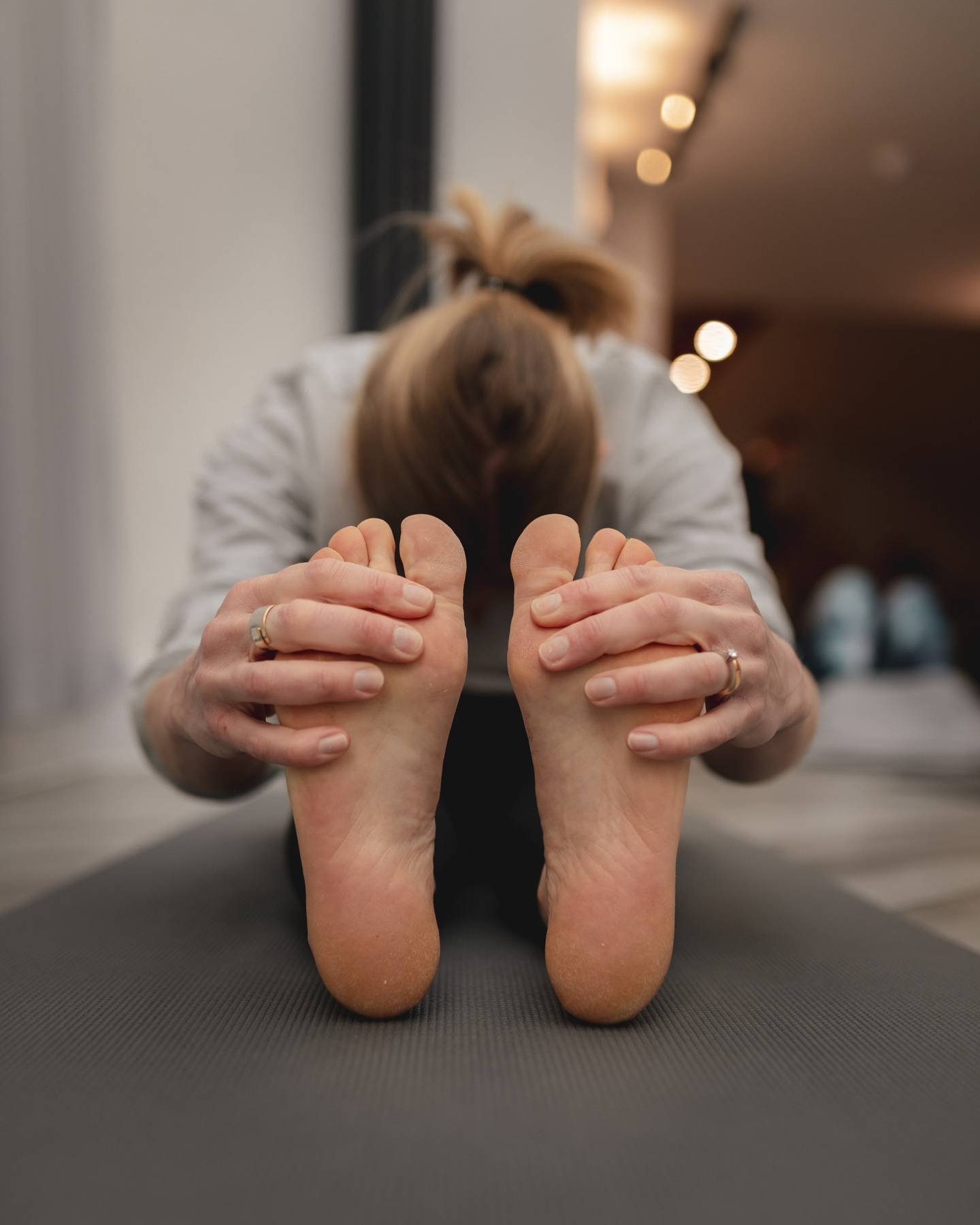 A person attending a yoga class in a studio, wearing a gray top, performing a forward bend pose with hands on feet, at The Basement Snug Yoga Studio in Kenilworth.