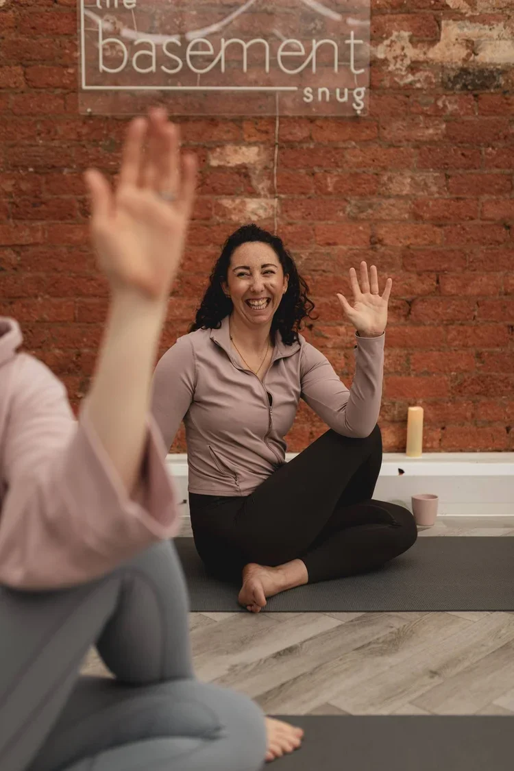 A woman sitting on a yoga mat with her legs crossed, smiling and raising her hands, during a yoga class at The Basement Snug Yoga Studio.