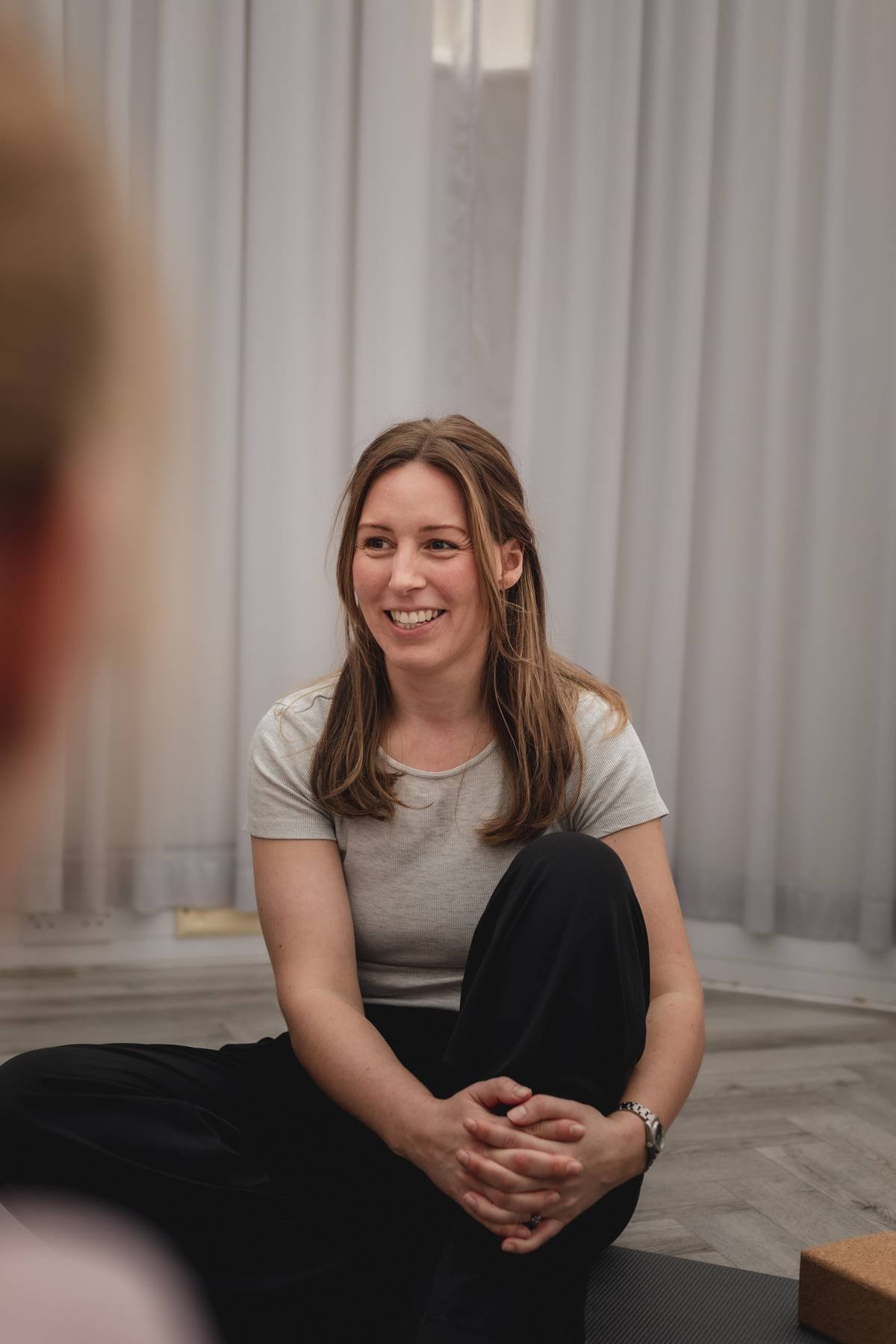 A woman sitting cross-legged on the floor, smiling and engaging in conversation at a Kenilworth yoga studio class.
