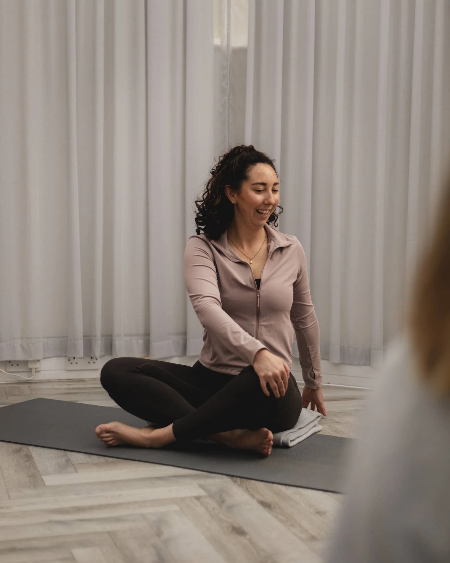 Woman in a pink athletic jacket and black leggings sitting cross-legged on a yoga mat, smiling during a at a Kenilworth yoga studio class.