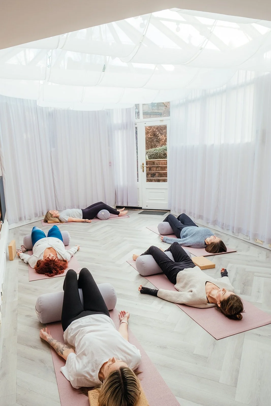 A group of people practicing yoga in a bright, airy room with white curtains and a door to the outside, at The Basement Snug Yoga Studio in Kenilworth.