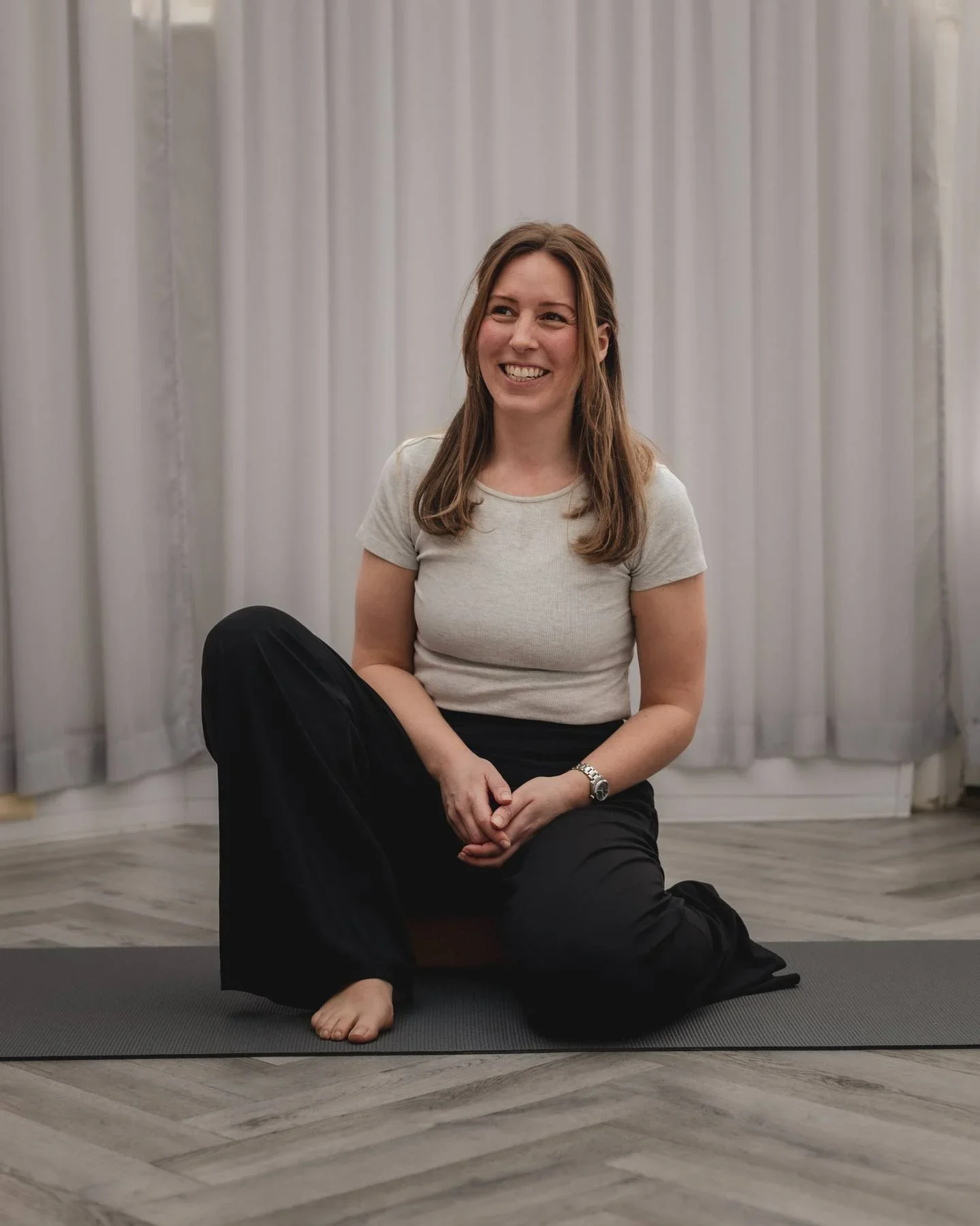 A yoga teacher wearing a light-coloured t-shirt and black pants, sitting on a yoga mat with her right leg bent and her left leg extended at The Basement Snug Yoga Studio in Kenilworth.