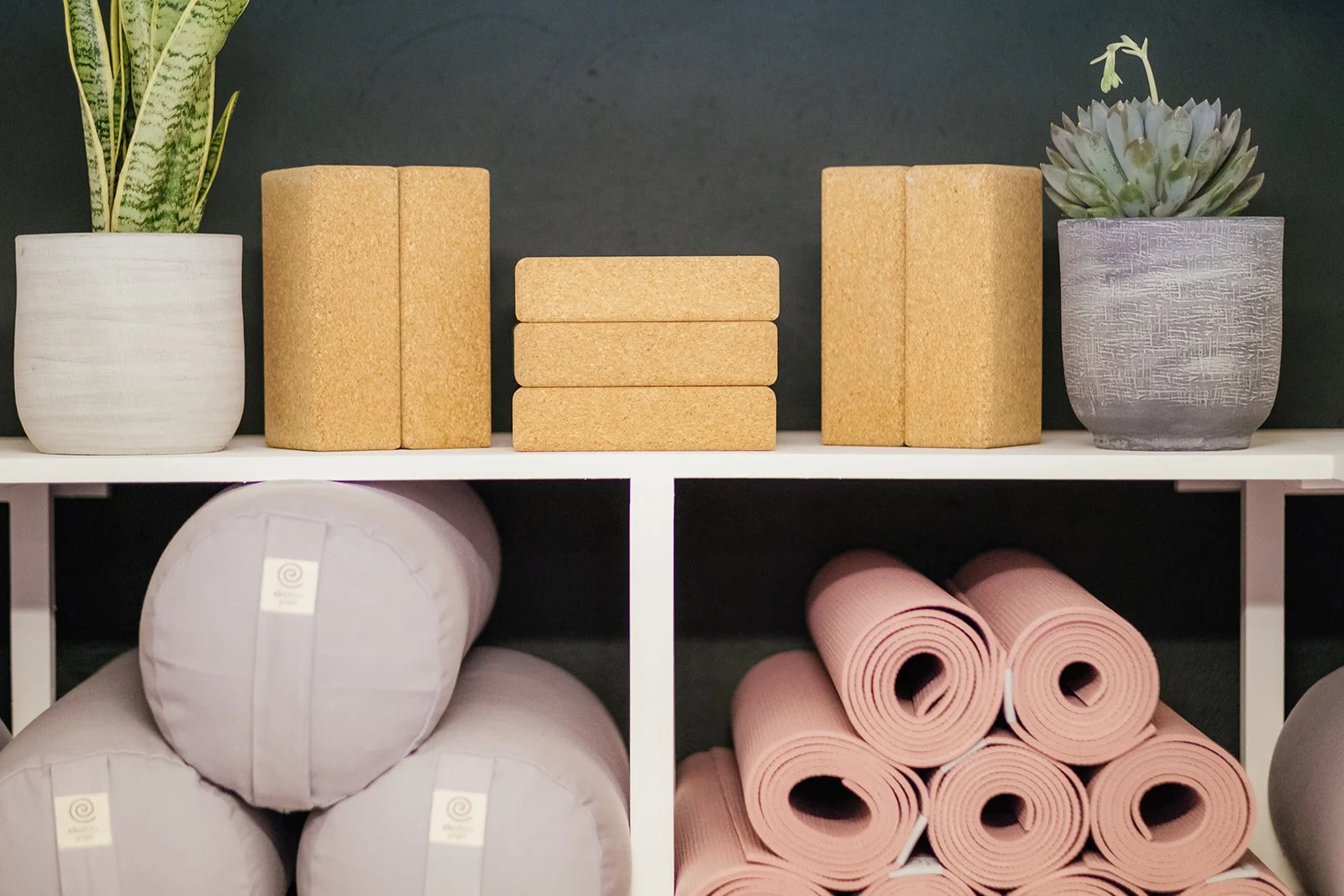 Organized yoga or fitness equipment on shelves, including potted succulents, cork blocks, pink yoga mats, and foam rollers at The Basement Snug Yoga Studio in Kenilworth.