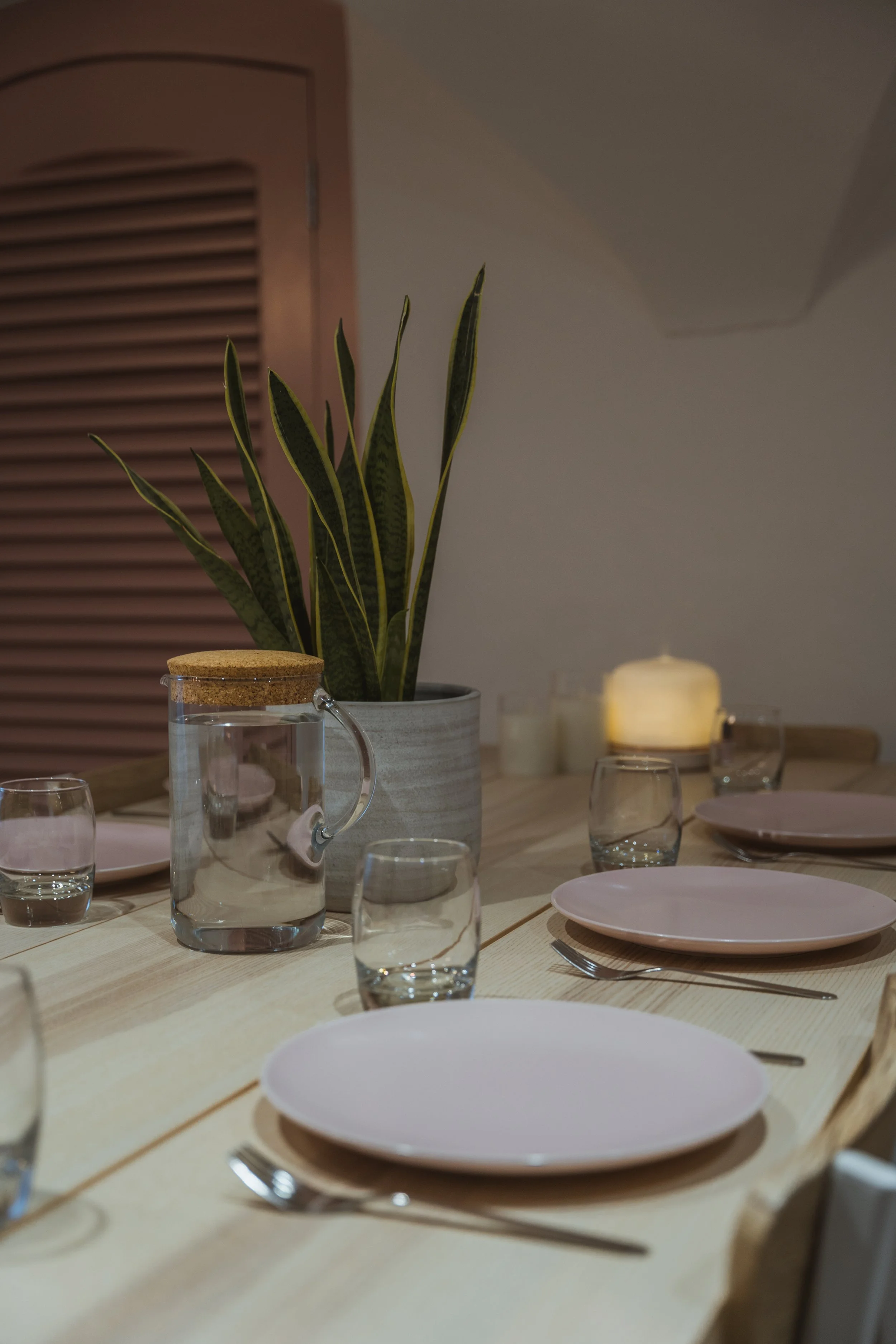 A dining table set with pink plates, clear glasses, silverware, a pitcher of water, at The Basement Snug Yoga Studio in Kenilworth.