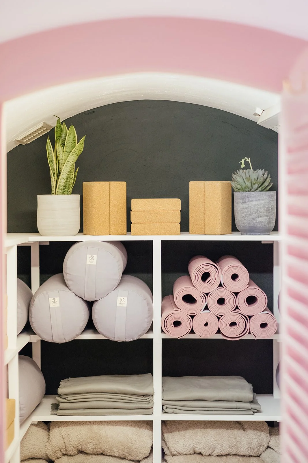 A closet storage shelf with yoga mats, blankets, and meditation cushions, with potted plants on top at The Basement Snug Yoga Studio in Kenilworth.