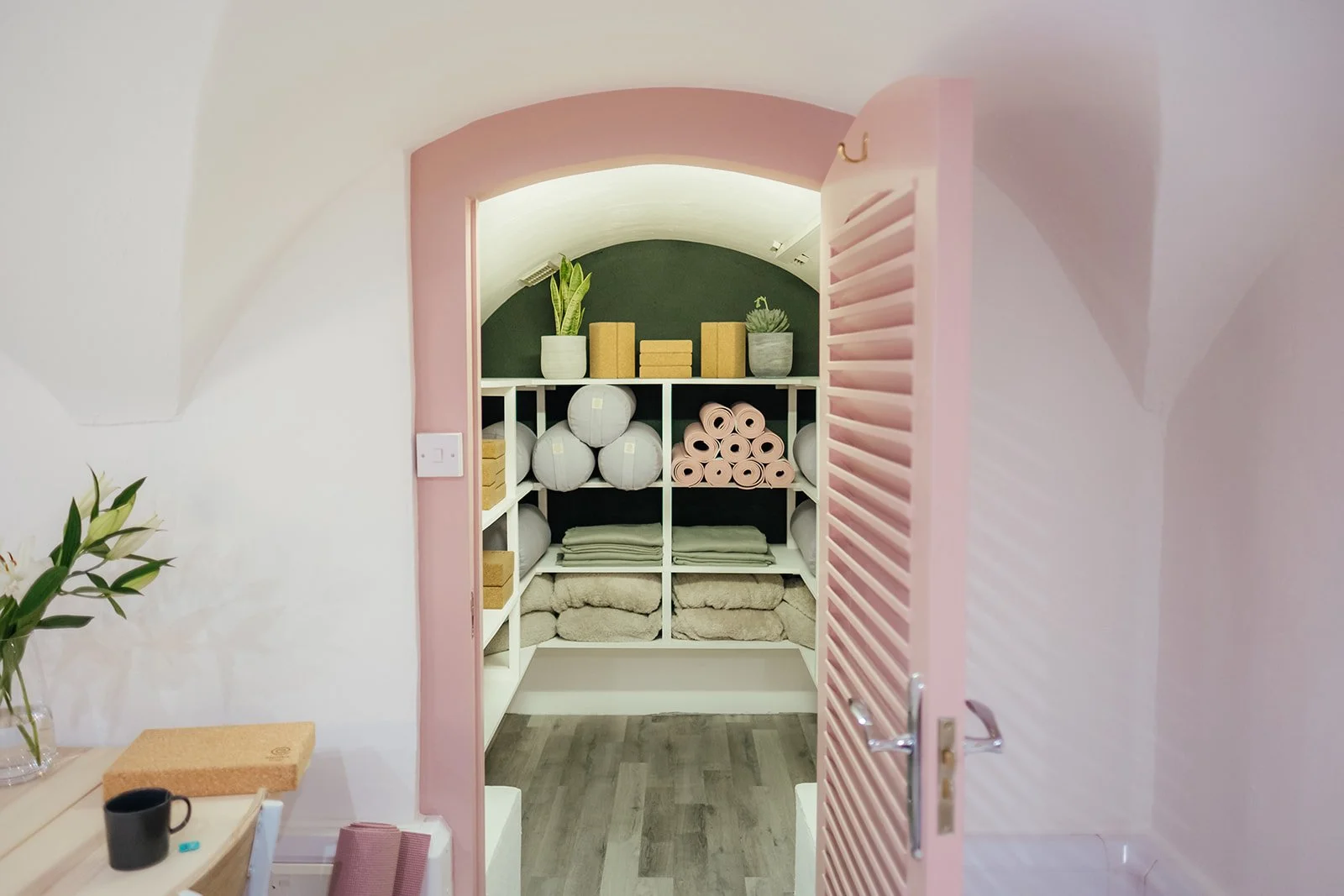 View of a cosy storage closet with shelves holding rolled towels, potted plants, and decorative boxes at The Basement Snug Yoga Studio in Kenilworth.
