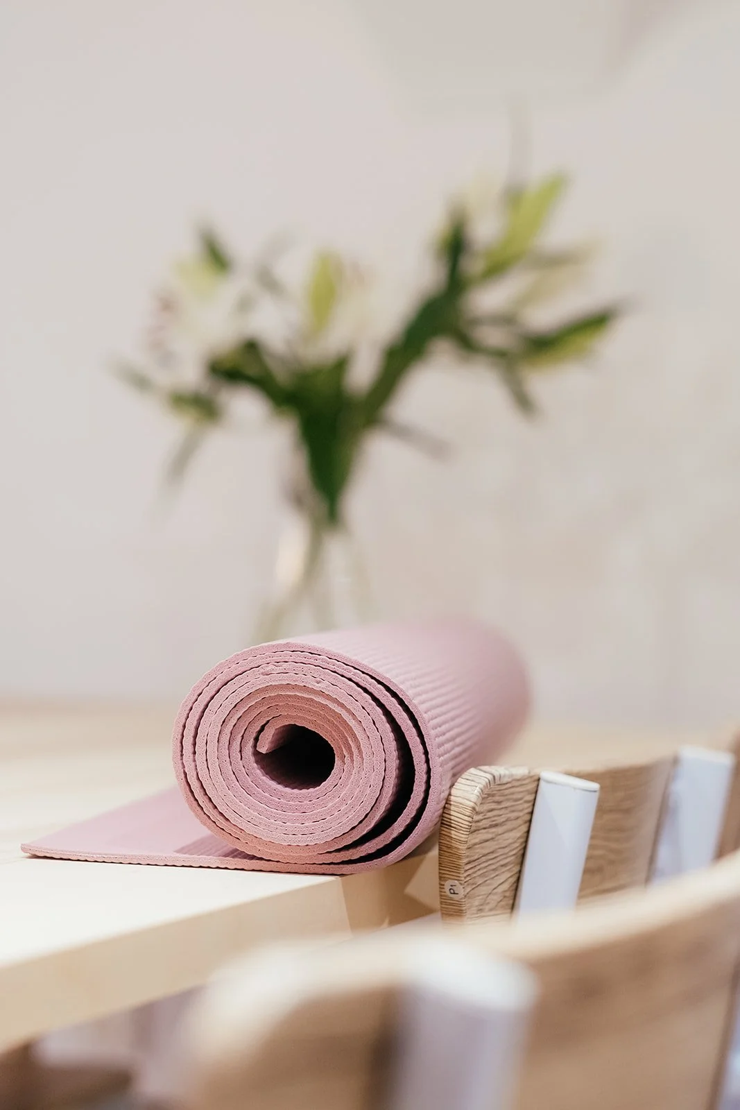 Pink rolled yoga mat on a wooden surface with a blurred vase of white flowers in the background at The Basement Snug Yoga Studio in Kenilworth.