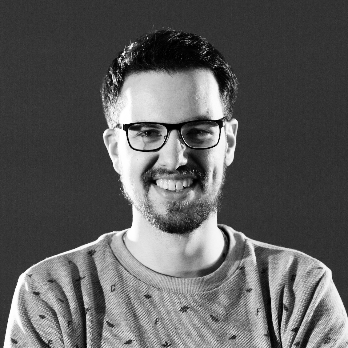Black and white portrait of a smiling man with glasses, a beard, and short hair, wearing a sweater with small patterns.