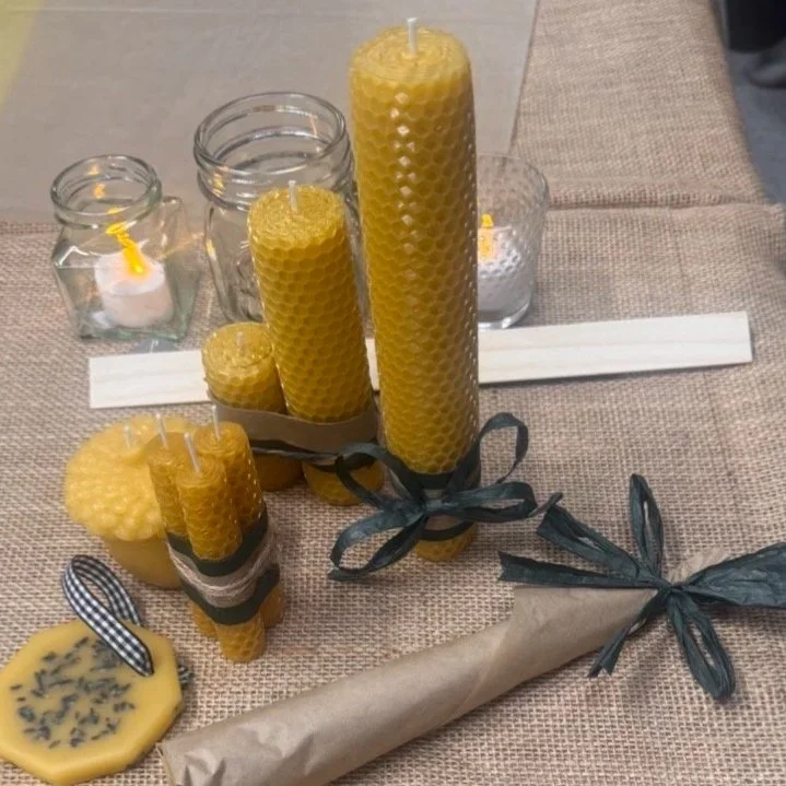 Various yellow beeswax candles of different sizes, some with black and white ribbon bows, placed on a beige tablecloth with candles in glass jars and a wrapped item.